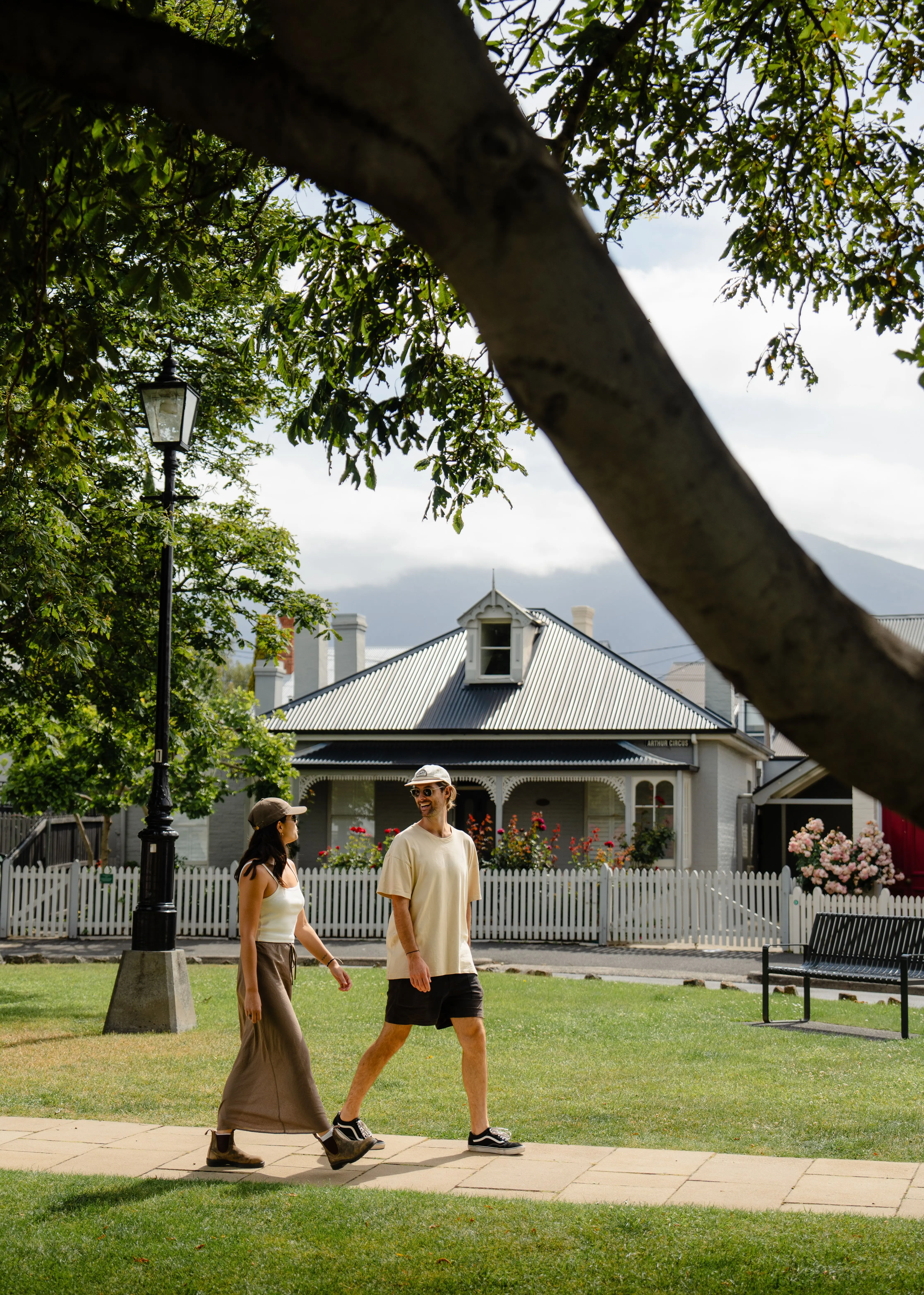 A man and woman in summer clothes walk past an old-fashioned lantern surrounded by grass and trees in a suburban street with white-picket fences and quaint cottages.