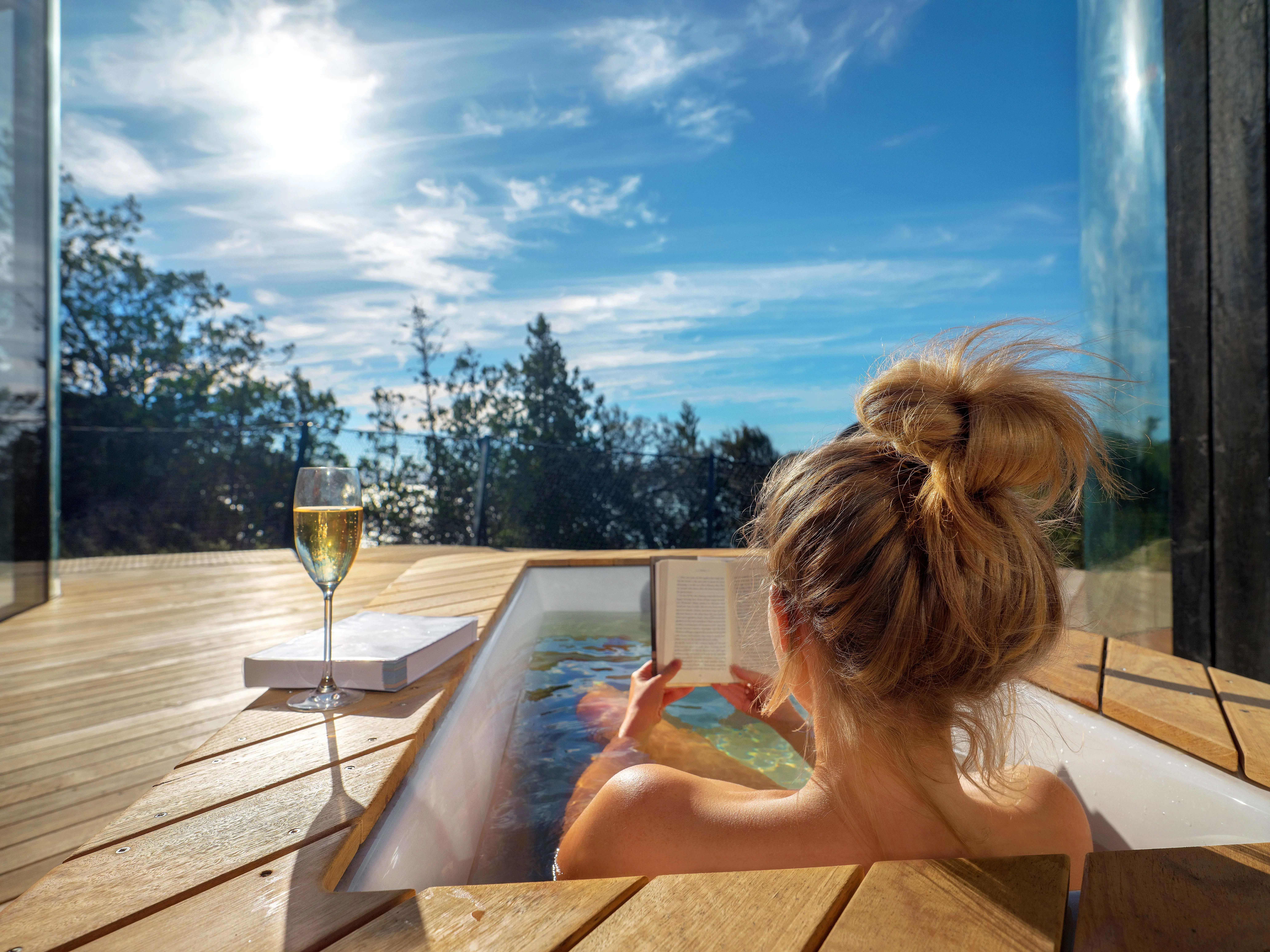 A young woman reclines in an outdoor bath overlooking forest and reads a book.