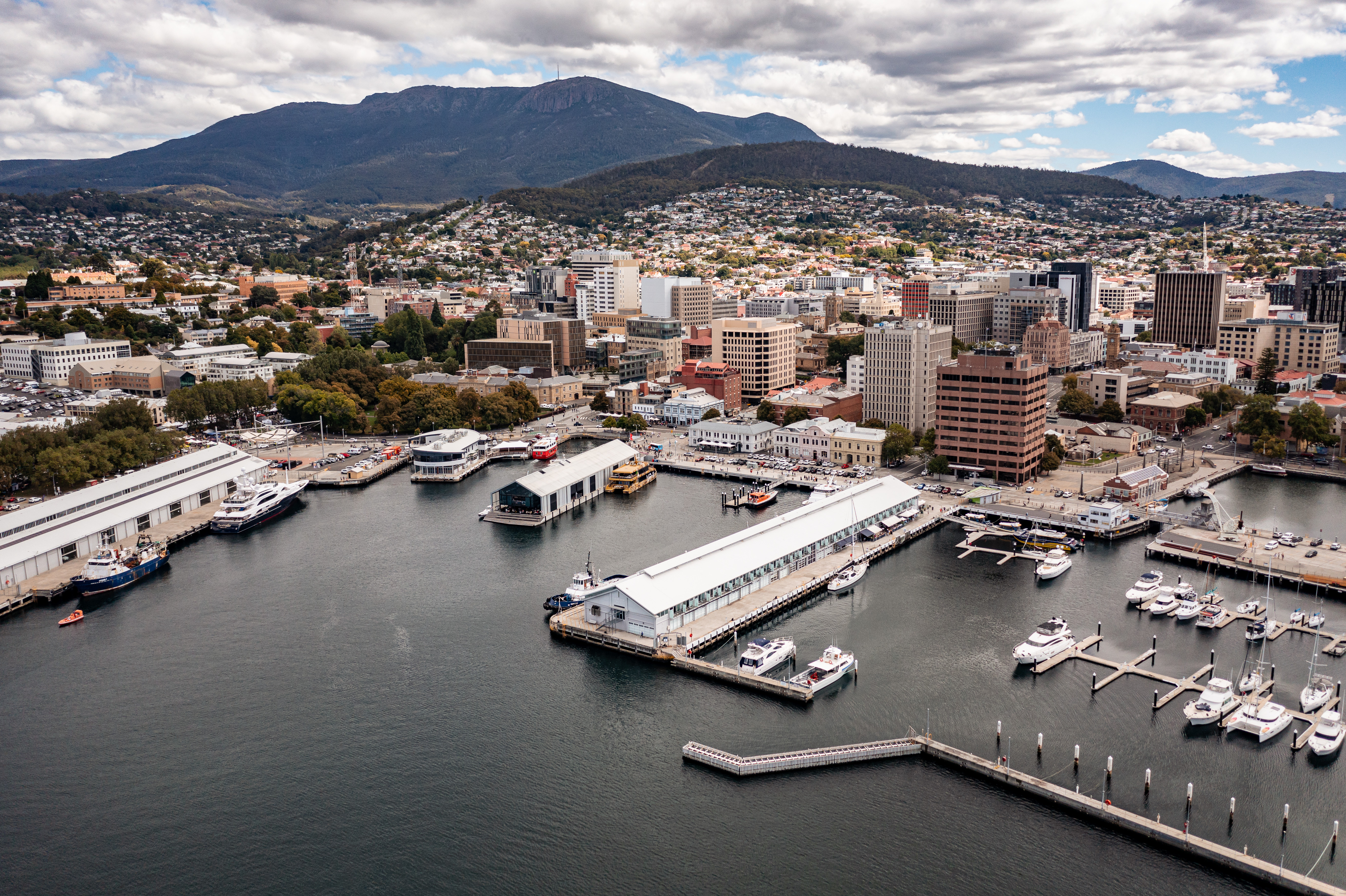An aerial view of Hobart's waterfront, with ships moored, long piers, the CBD buildings and beyond, a mountainous landscape.