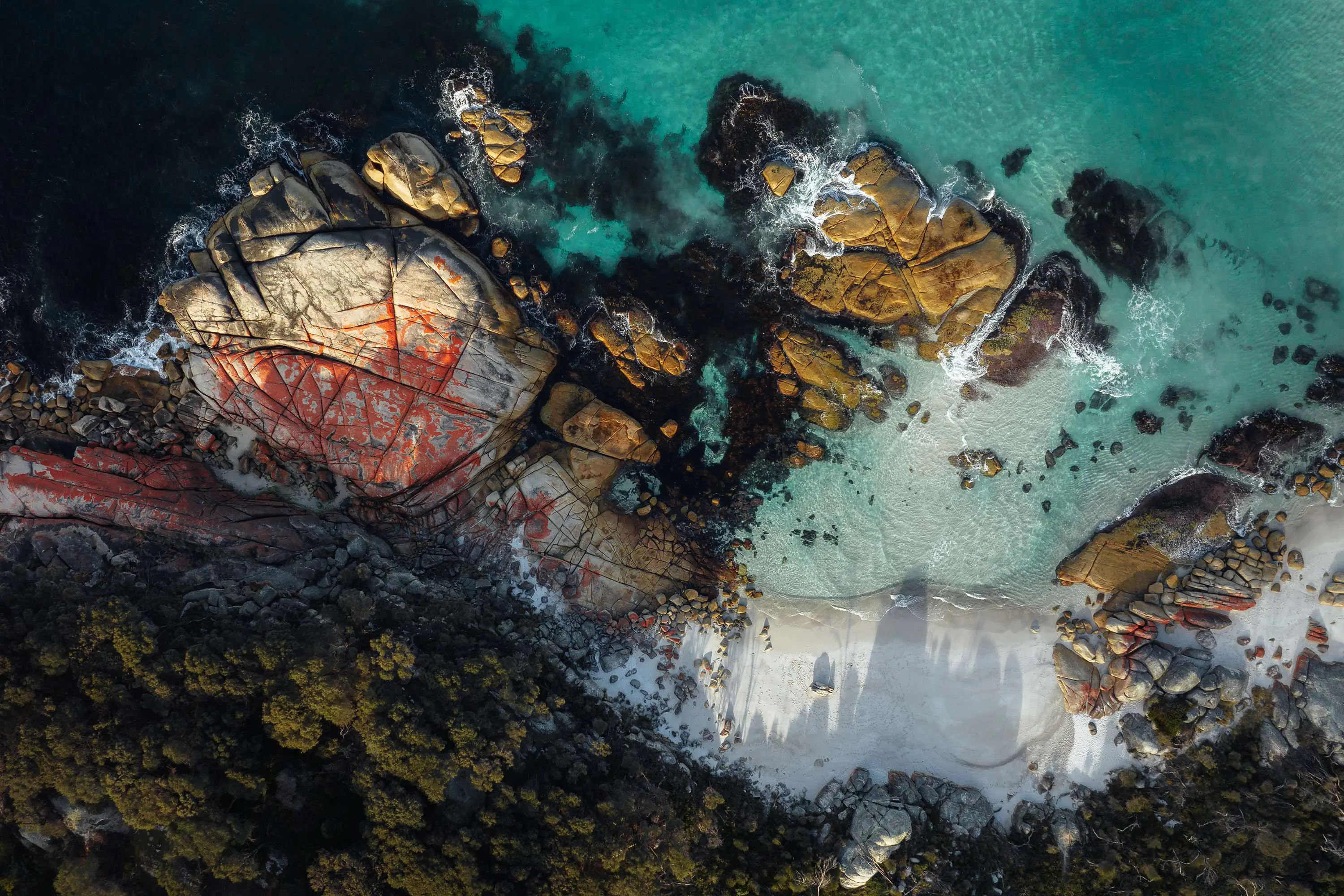 A top-down aerial view of a stunning beach. The water is turquoise and clear, huge boulders with orange lichen are scattered about and dark bush edges the white sand.