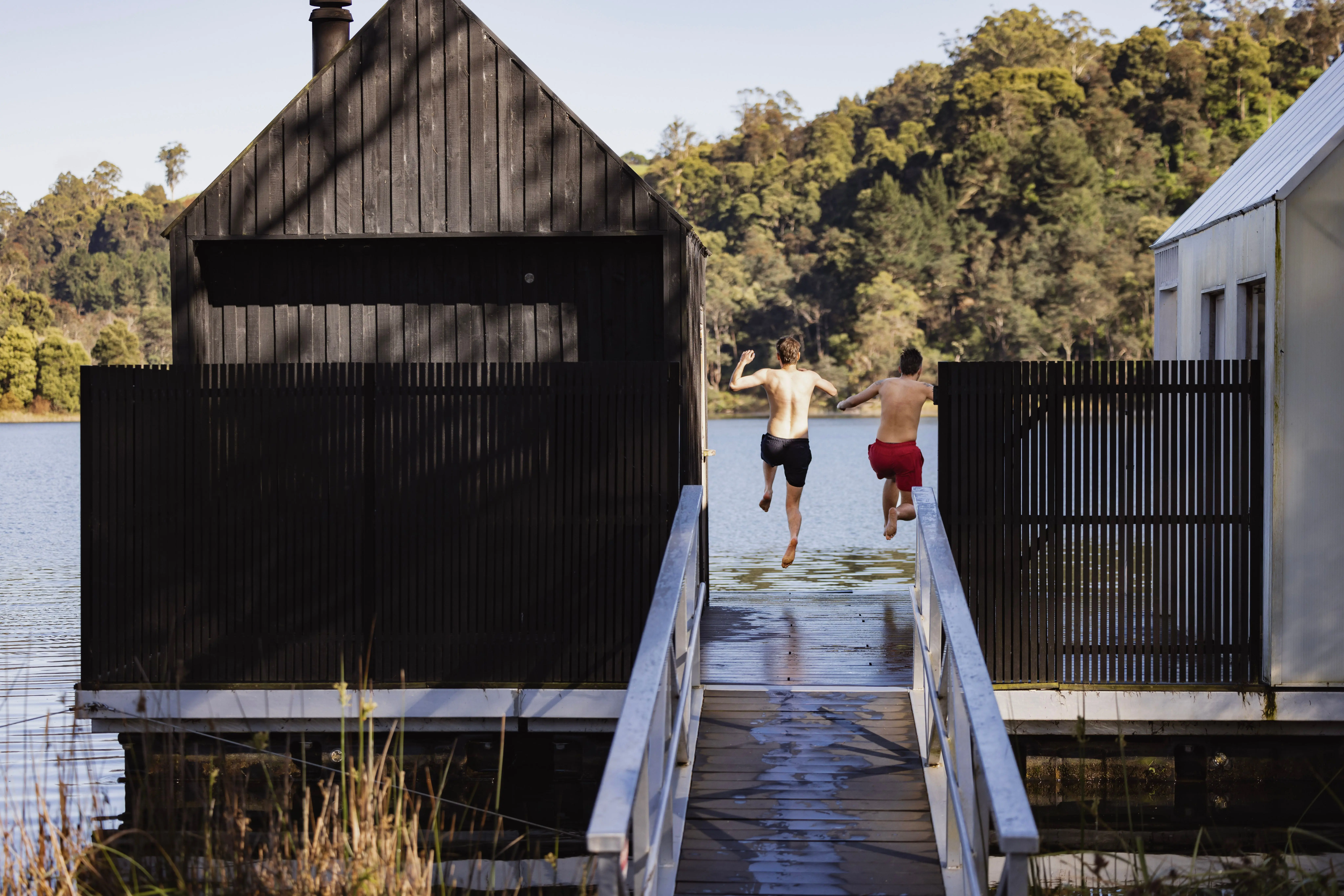 Two men jump off a pier between a black timber-clad building and a light building that float above a lake.