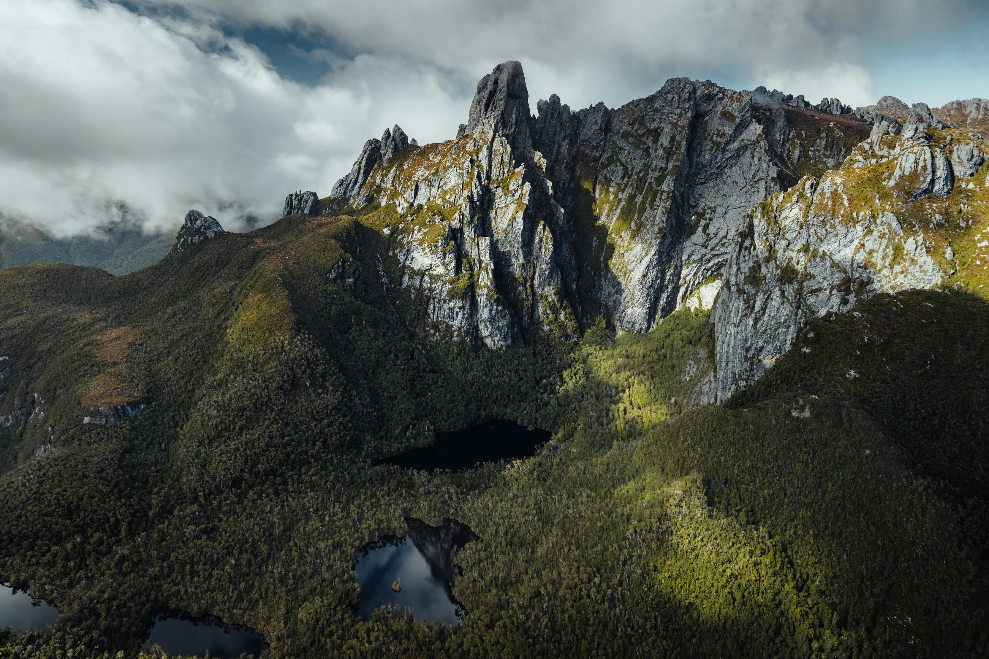 An aerial view of Federation Peak, a sharp, craggy mountain jutting out of thick dense bushy hills. Heavy cloud rolls overhead and still lakes reflect the sky amongst the trees below. 