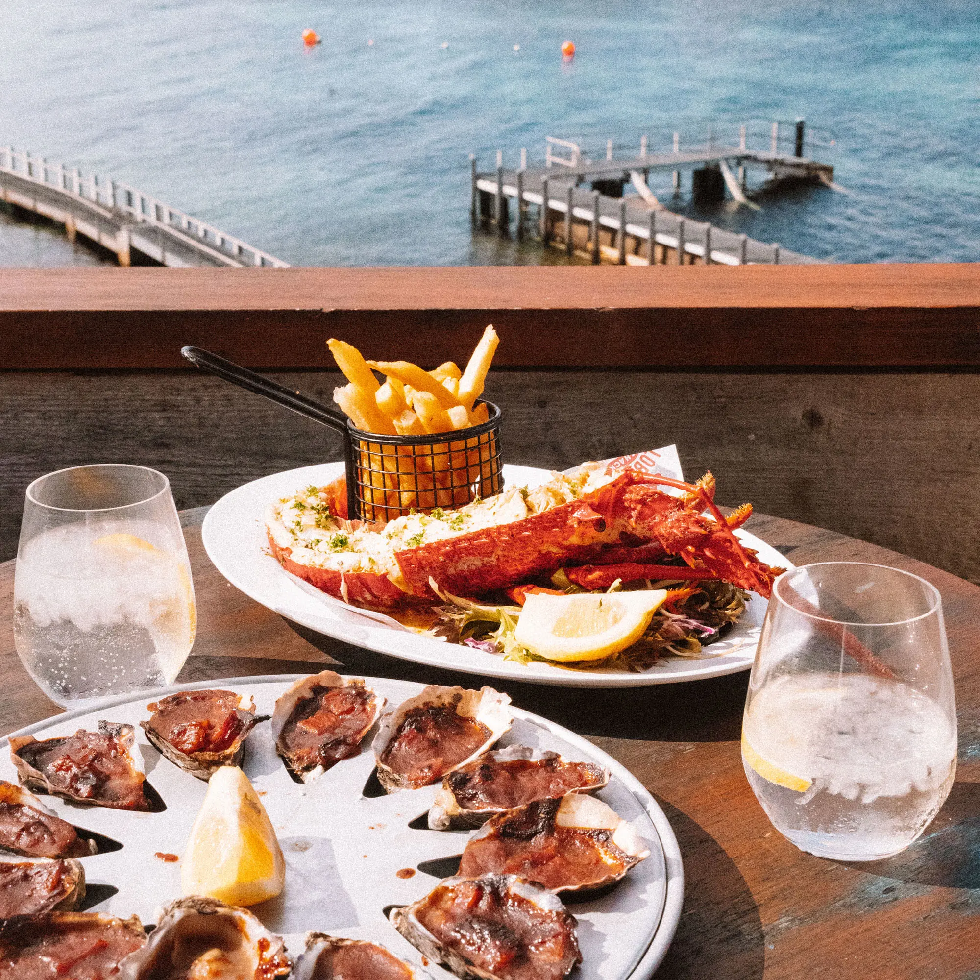 A table, overlooking a calm bay with wooden jetties, displays plates of oysters and whole cooked lobster with chips, lemon and water glasses.