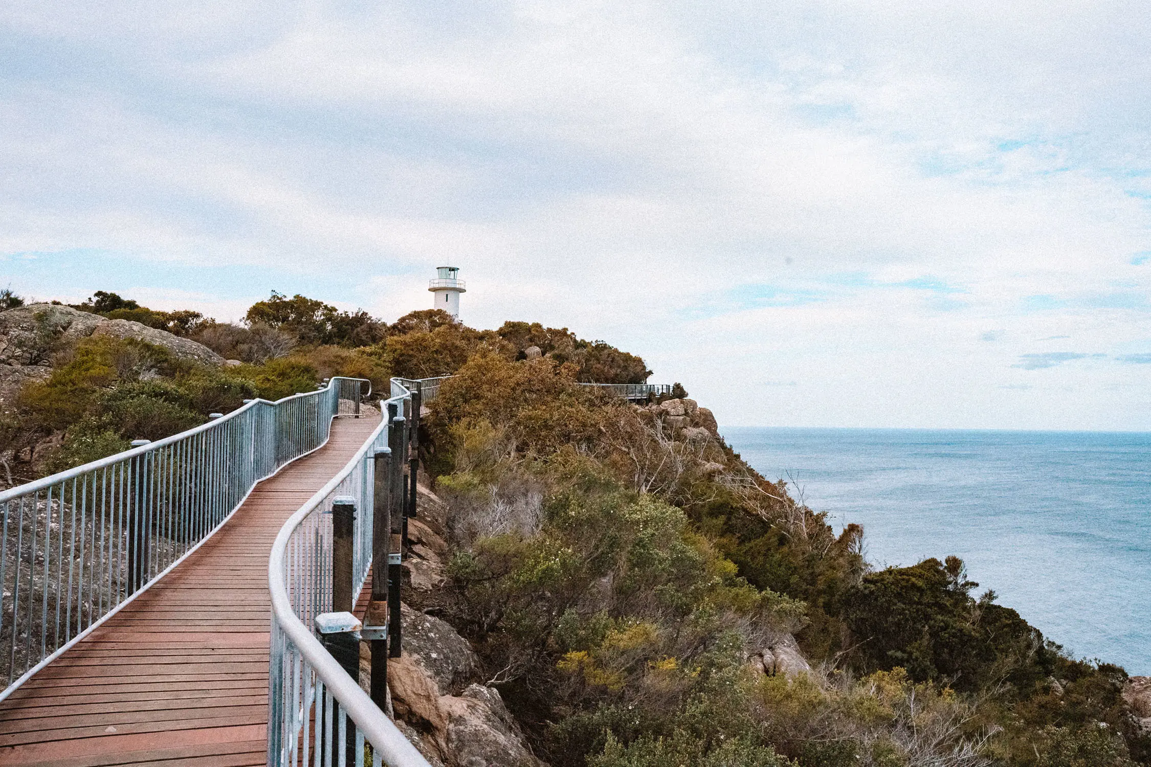 A curved timber boardwalk with metal railings follows rocky coastal headland covered in native vegetation. A lighthouse is visible at the highest point.