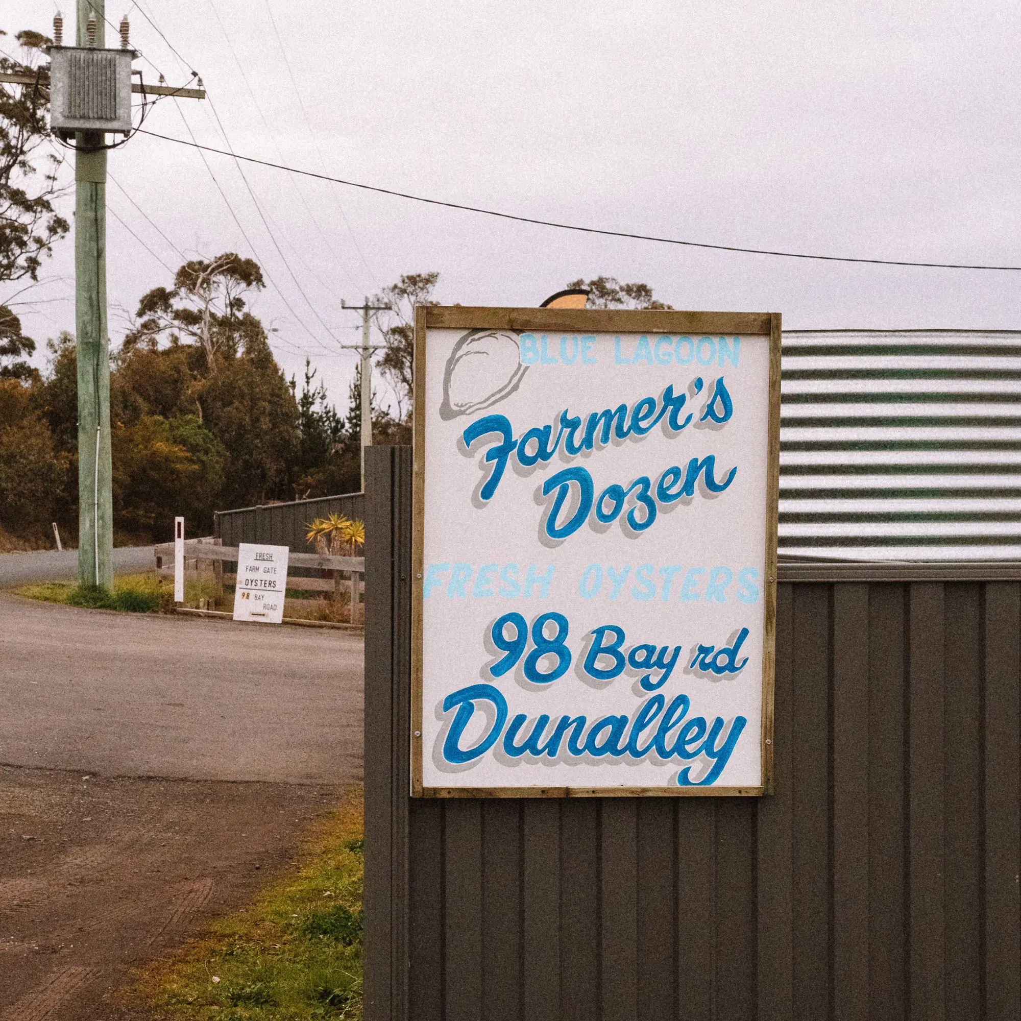 A hand-painted white sign reading "Farmer's Dozen" and "98 Bay Rd Dunalley" is mounted on a weathered wooden fence.