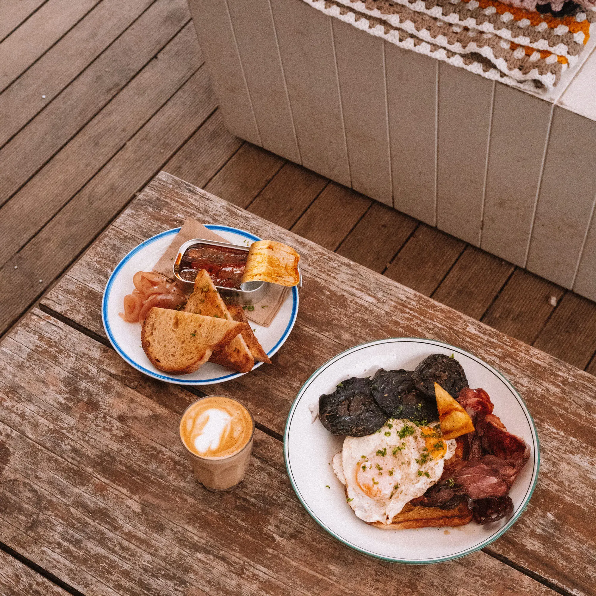 Two white plates sit on weathered timber decking, holding freshly cooked breakfast. A glass of juice sits nearby.