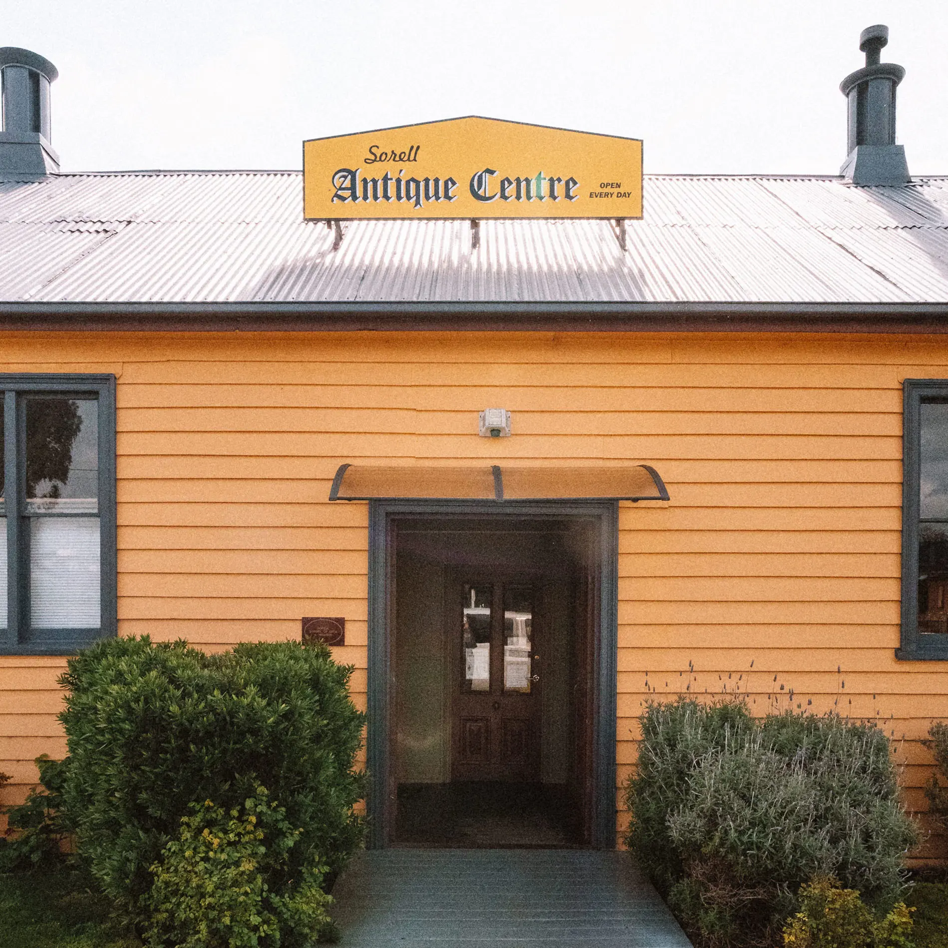 A honey-colored wooden building with a dark entrance door sits behind landscaped shrubs on a graveled pathway. A sign on the roof says "Sorell Antique Centre".