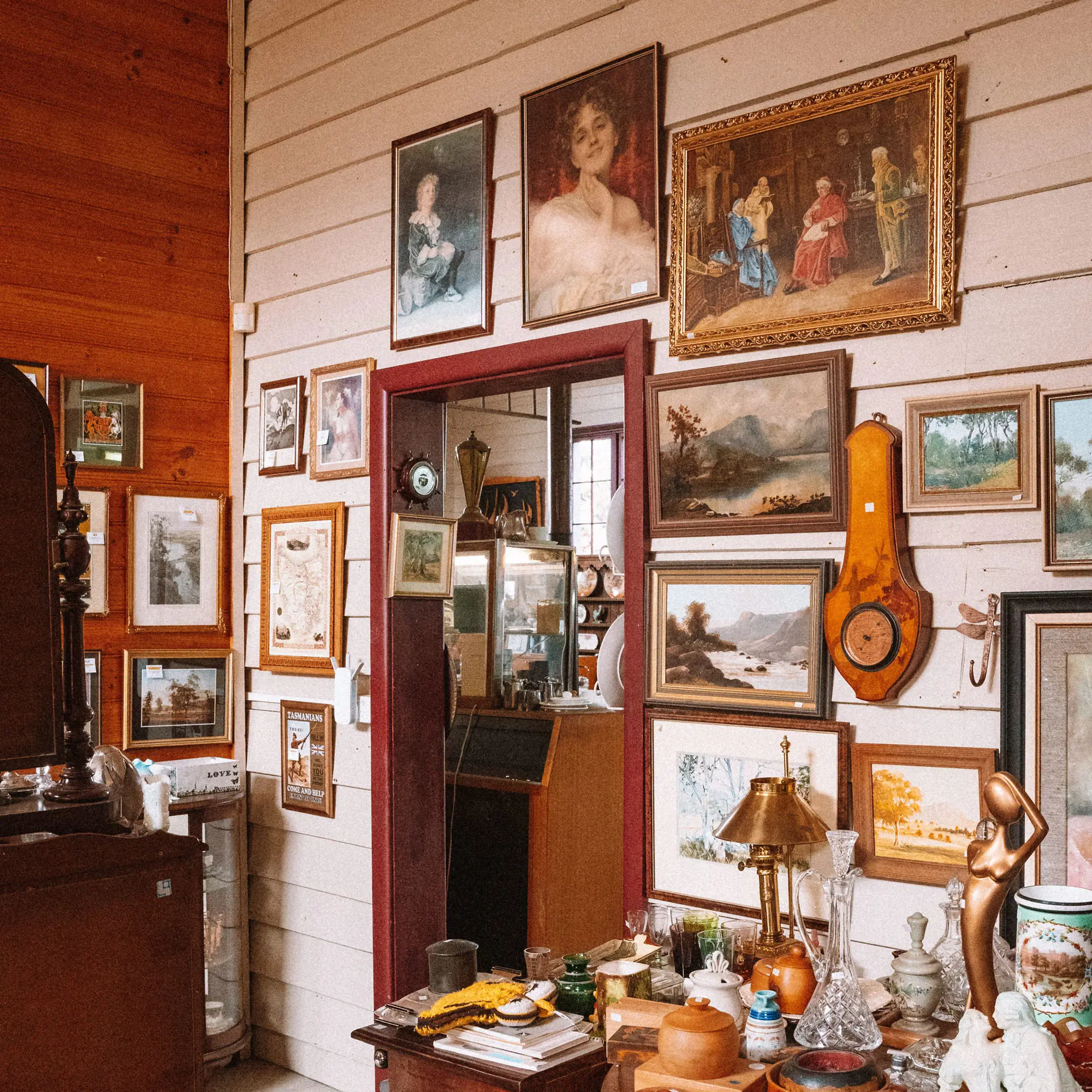 The corner of an antique shop, with the weatherboard walls covered in paintings and photos, and many curios atop the sideboard.