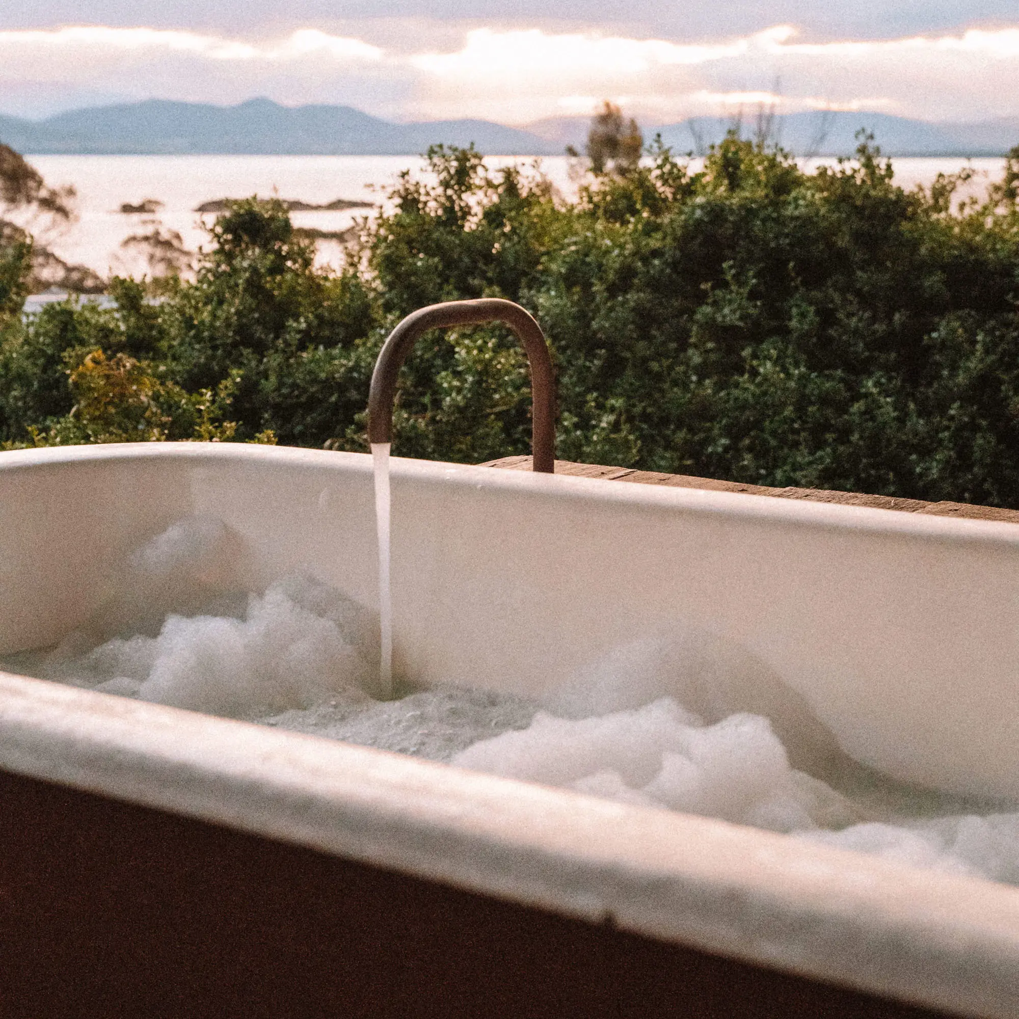A freestanding red and white bath, filling with bubbles, sits on a timber deck, overlooking the water and bush under a pale sky.