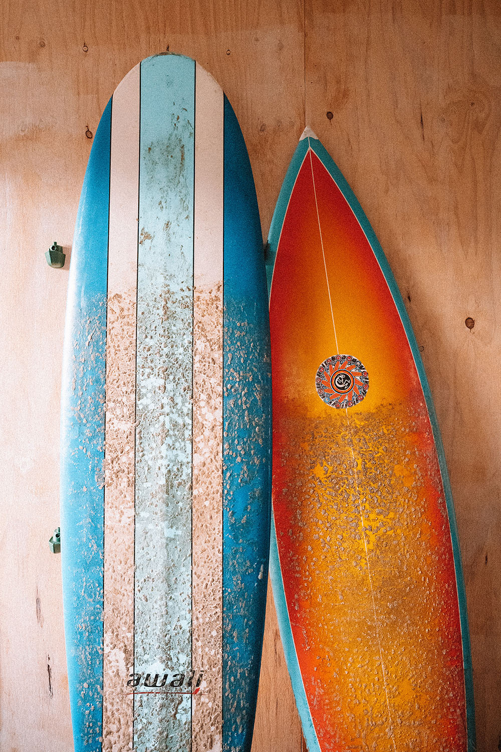 Two worn surfboards, colourful and dotted with wax, lean up against a light wooden-clad wall.