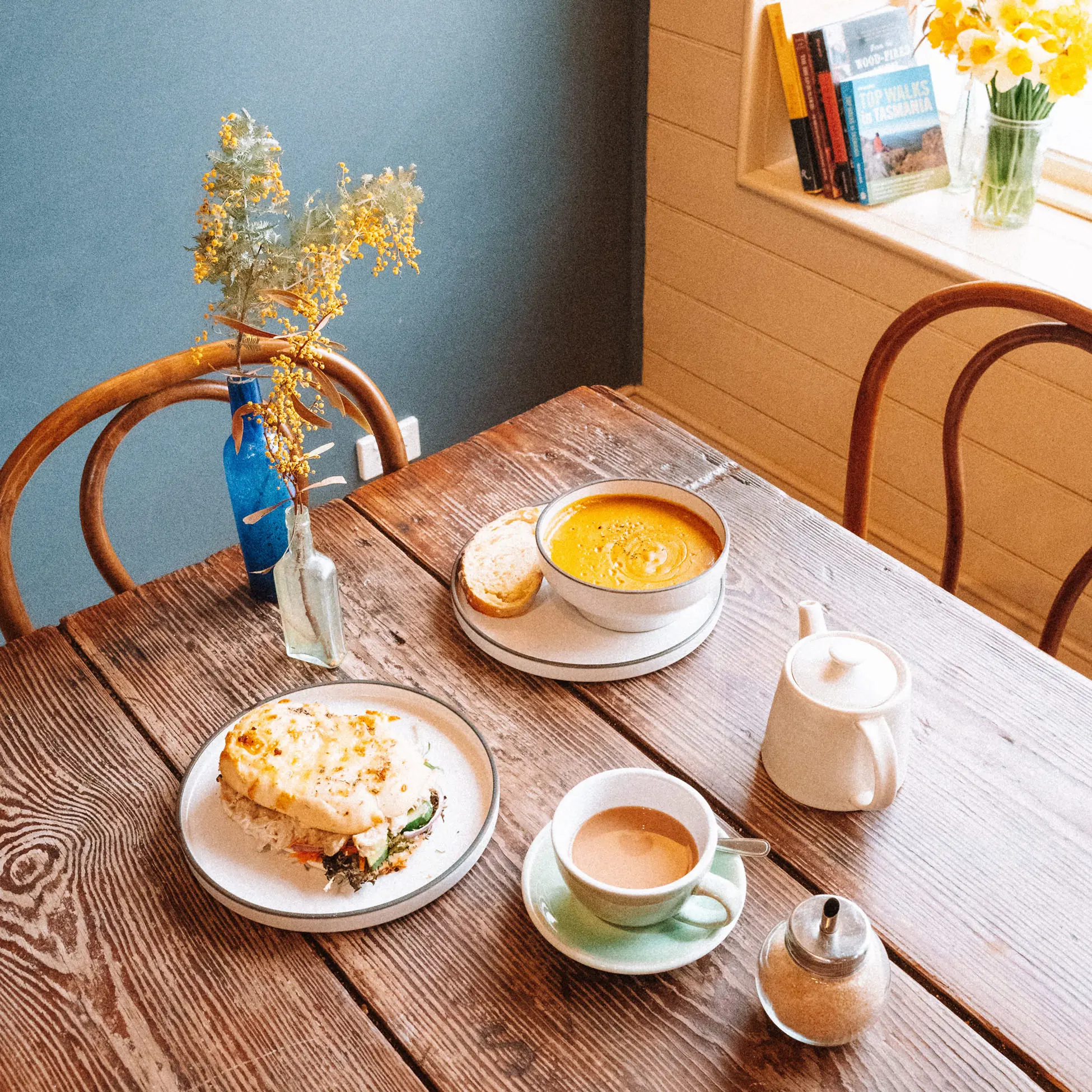 Hearty plates of soup, a toastie and a pot of tea sit on a wooden table in a brightly-lit cafe.