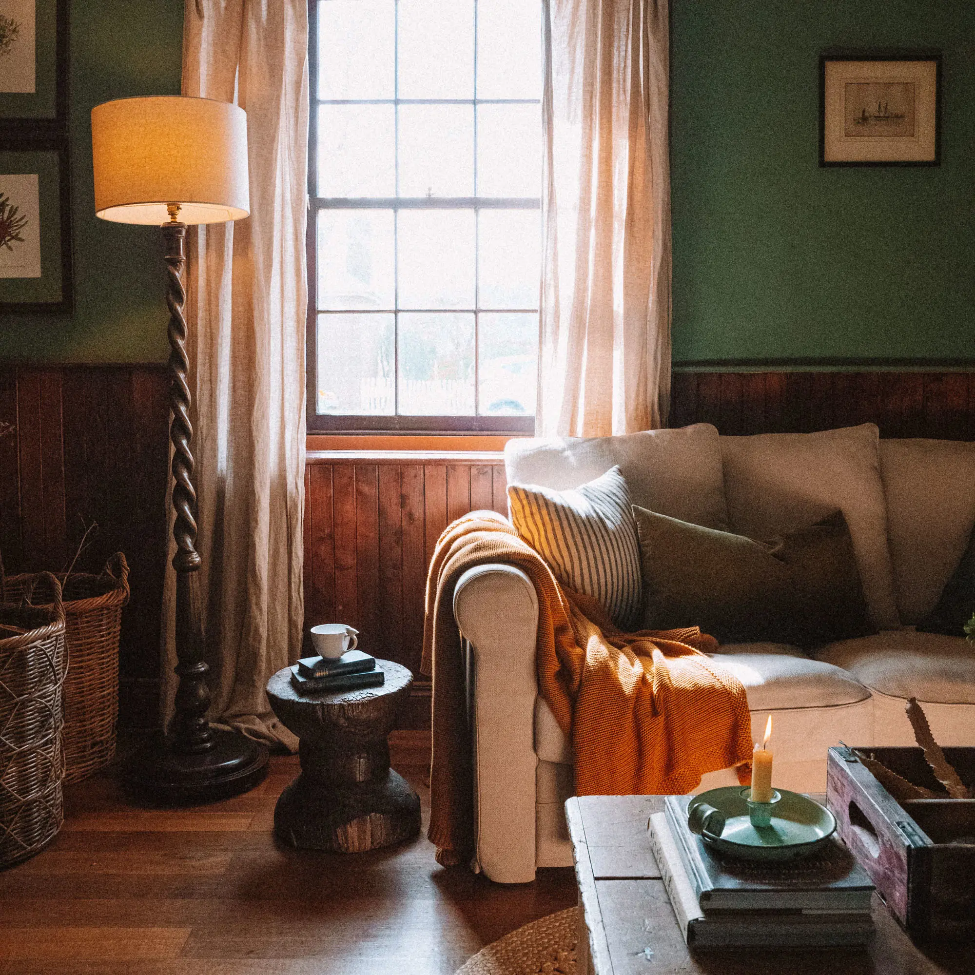 A cosy living room, artfully styled with throws and pillows on the cream couch, candles lit and the sunlight streaming through the sash window.