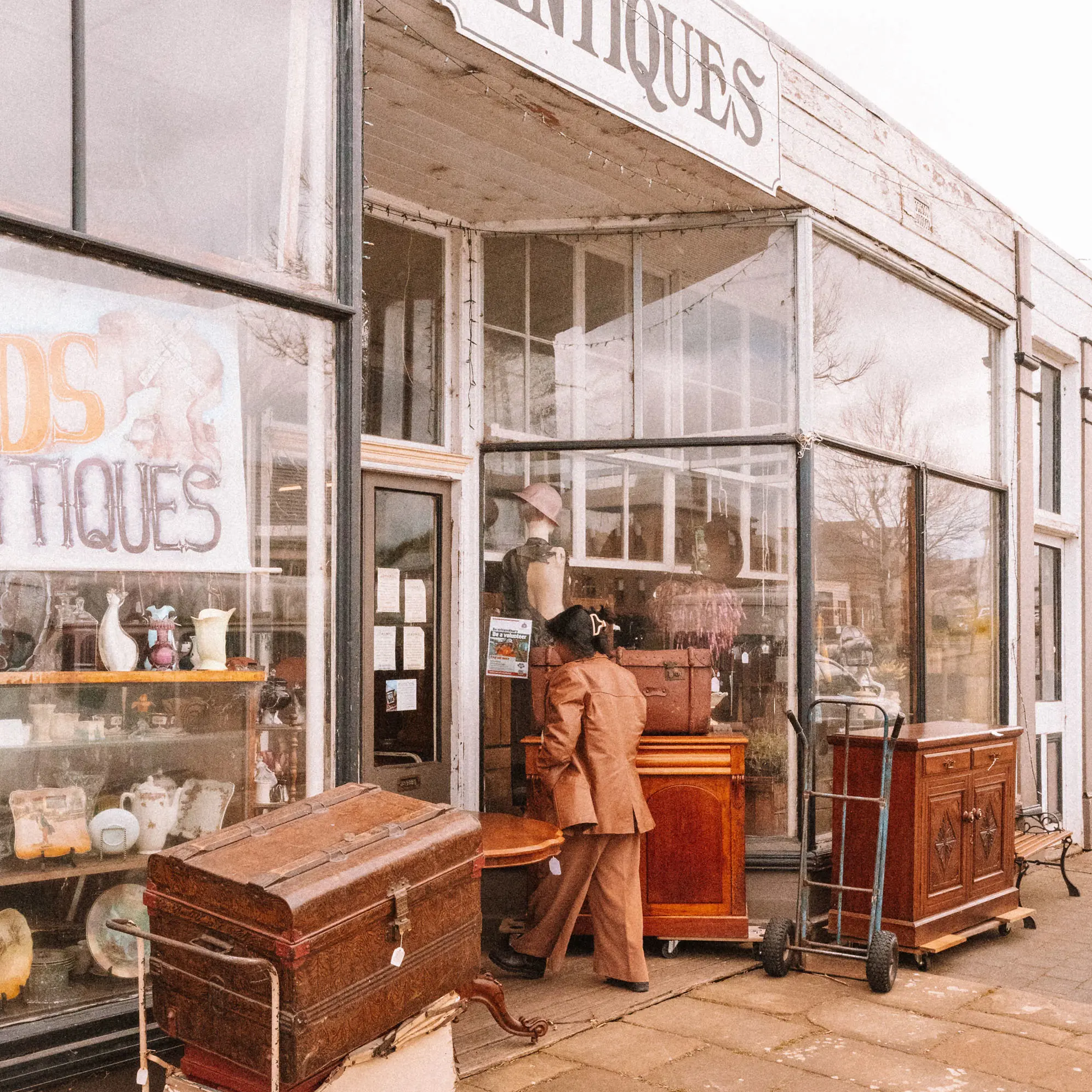 A woman in a brown suit walks through the doors of a weathered antique shop, with beautiful heritage furniture on display outside.