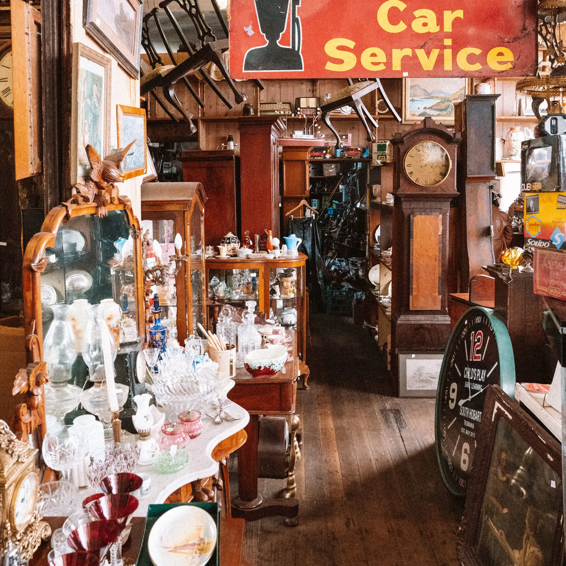 China, glasses and other vintage bric-a-brac line the shelves and furniture inside an antique shop. Chairs hang from the ceiling and a faded red and yellow sign says 'Car Service'.