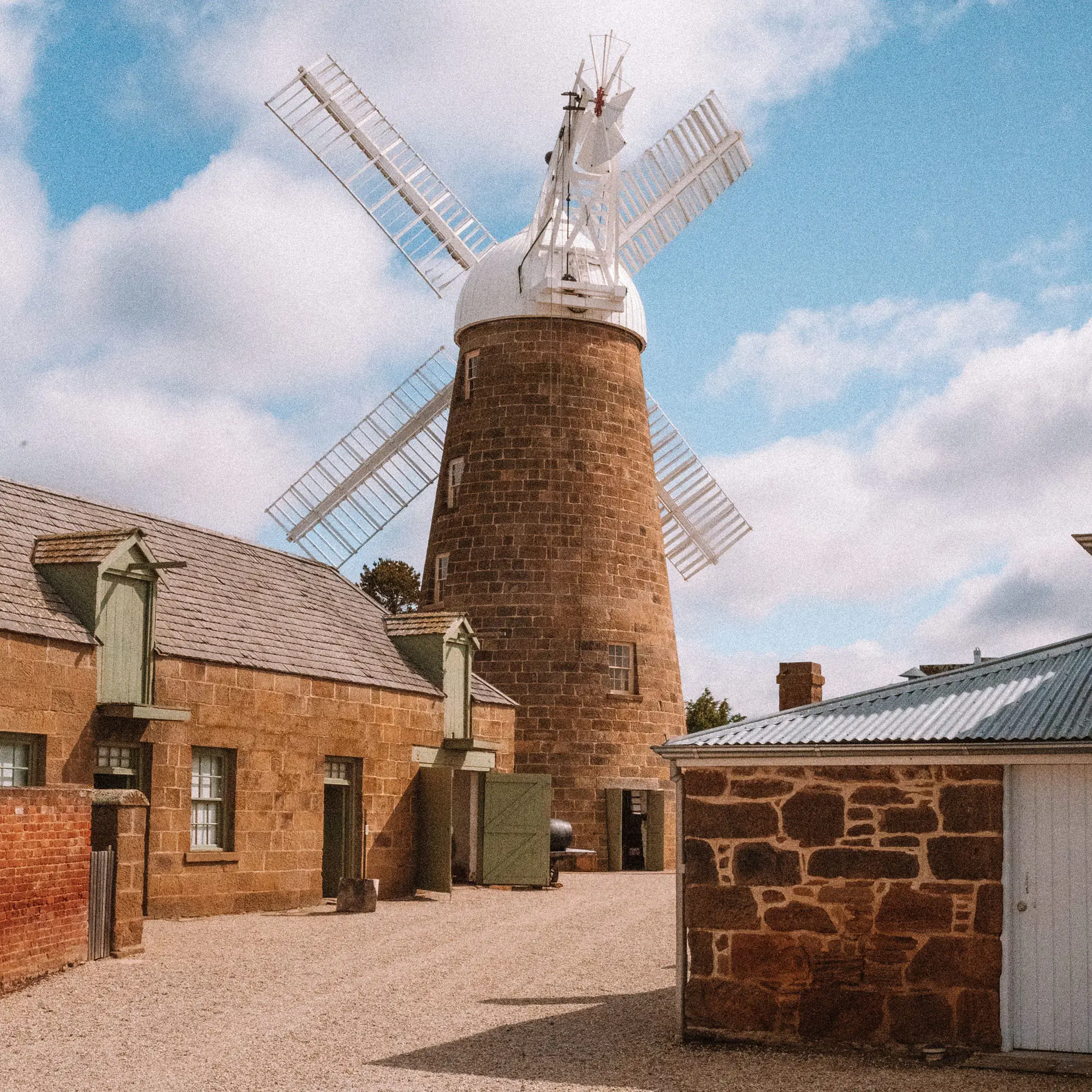 A historic windmill stands tall above a courtyard precinct of cottages and buildings with raked gravel ground.