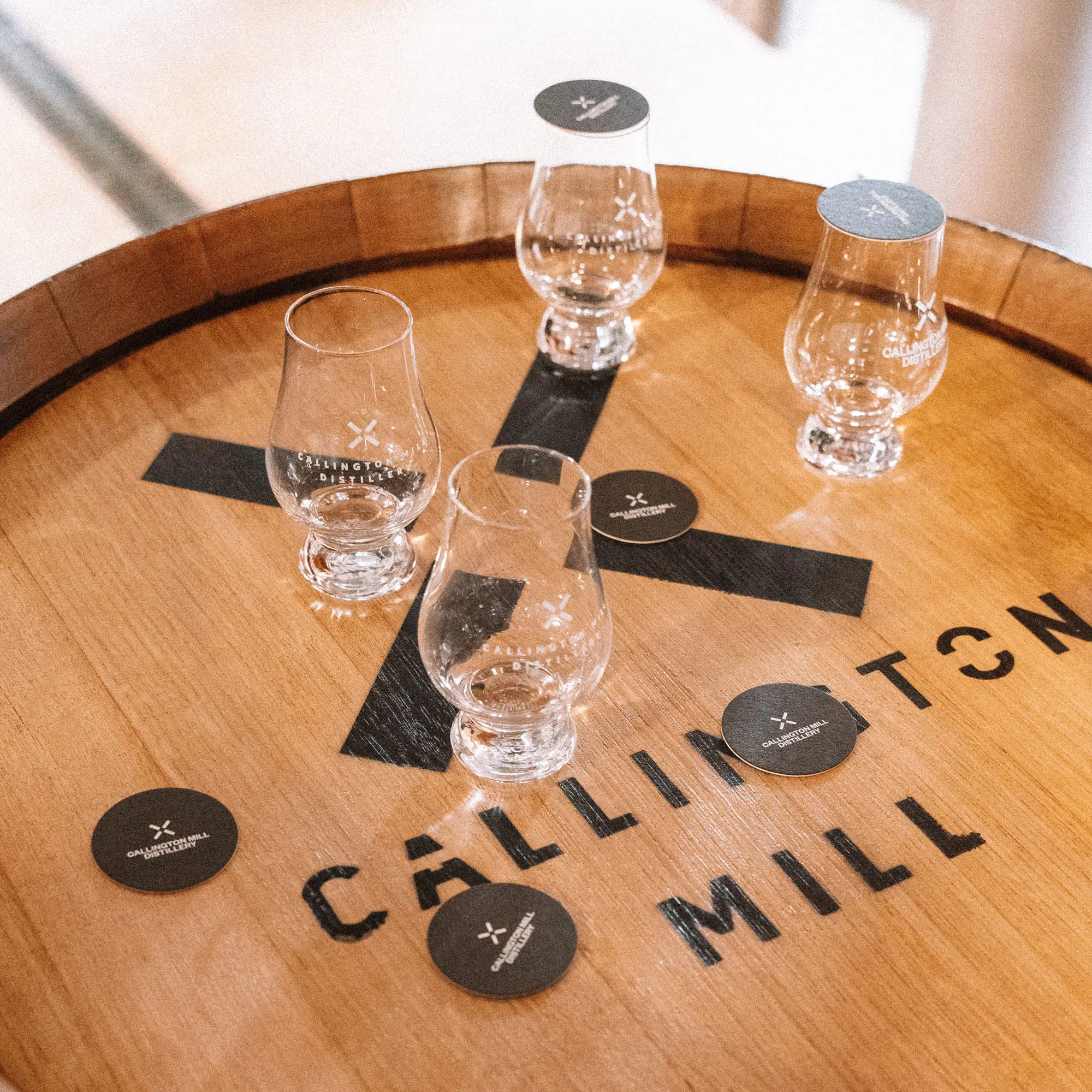 Four whisky glasses sit on top of a barrel, with a large X logo and the words 'Callington Mill' burned into the wood.