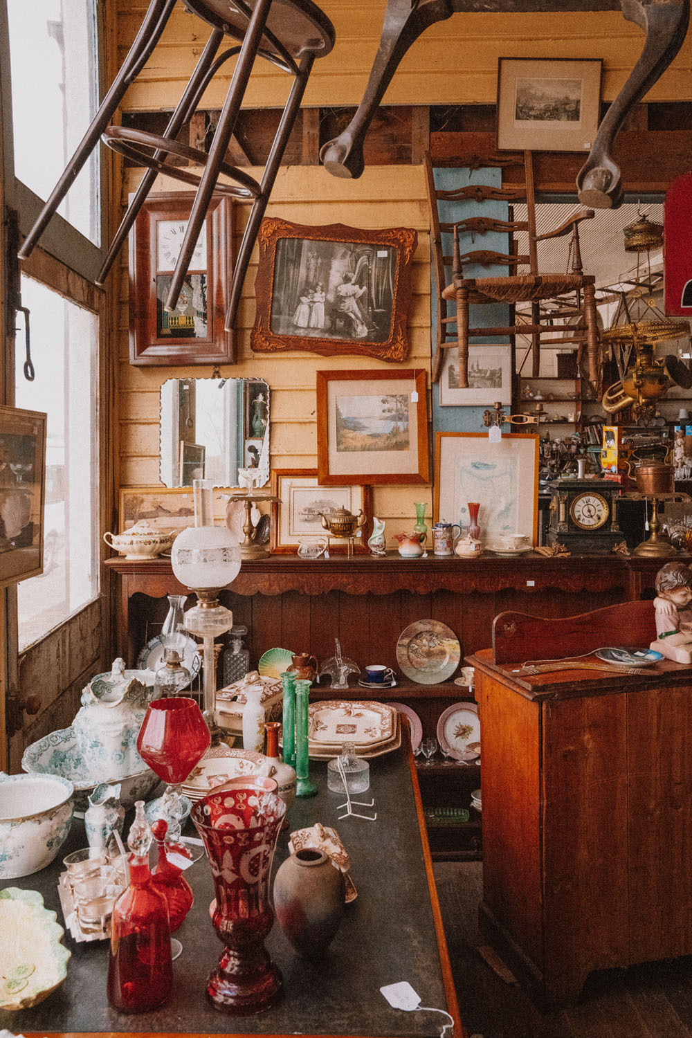 The interior of an antiques shop. The weatherboard wall is covered in hanging pictures and mirrors, chairs are suspended at angles from the ceiling, and a wooden sideboard is covered in treasures.
