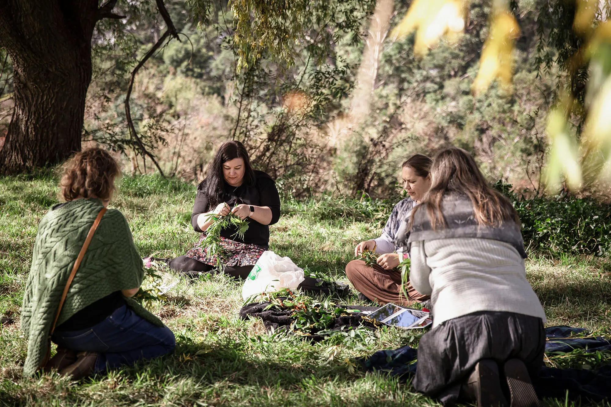 Four women sit on the grass under some trees, working on hand-weaving leaves, vines and other foliage.