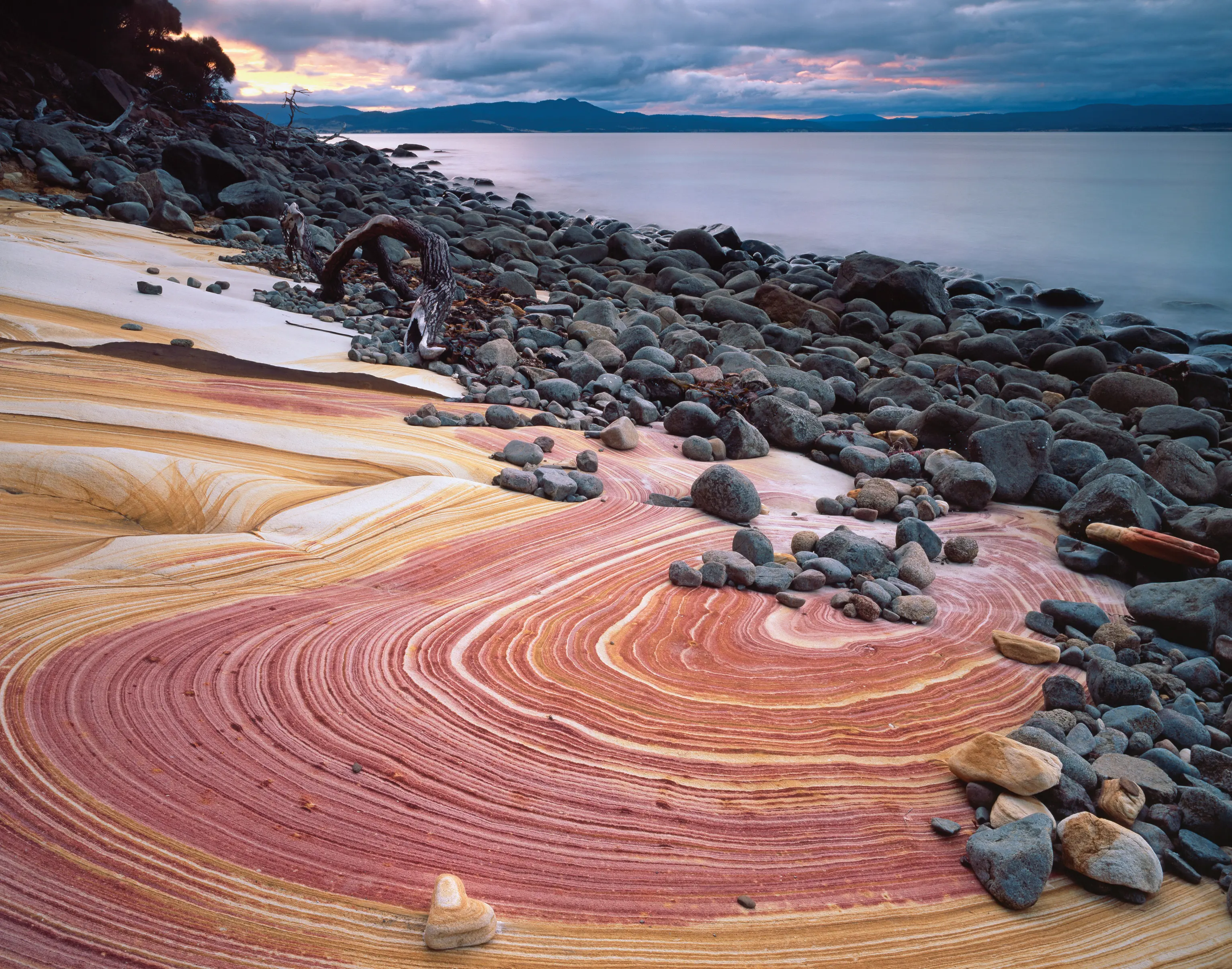 A striking rock formation on a rocky shoreline. The ground looks like it has been painted in red and orange swirls.