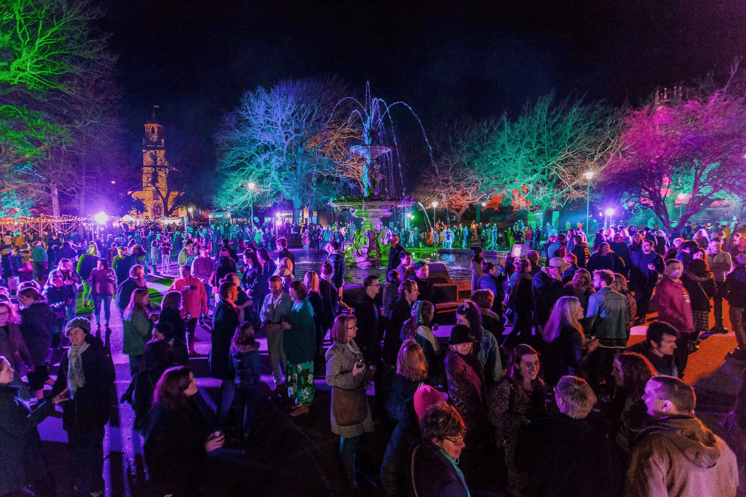 A crowd of people in an outdoor park at night. The people and the bare branches of trees are lit up in shades of green, pink, blue and purple.