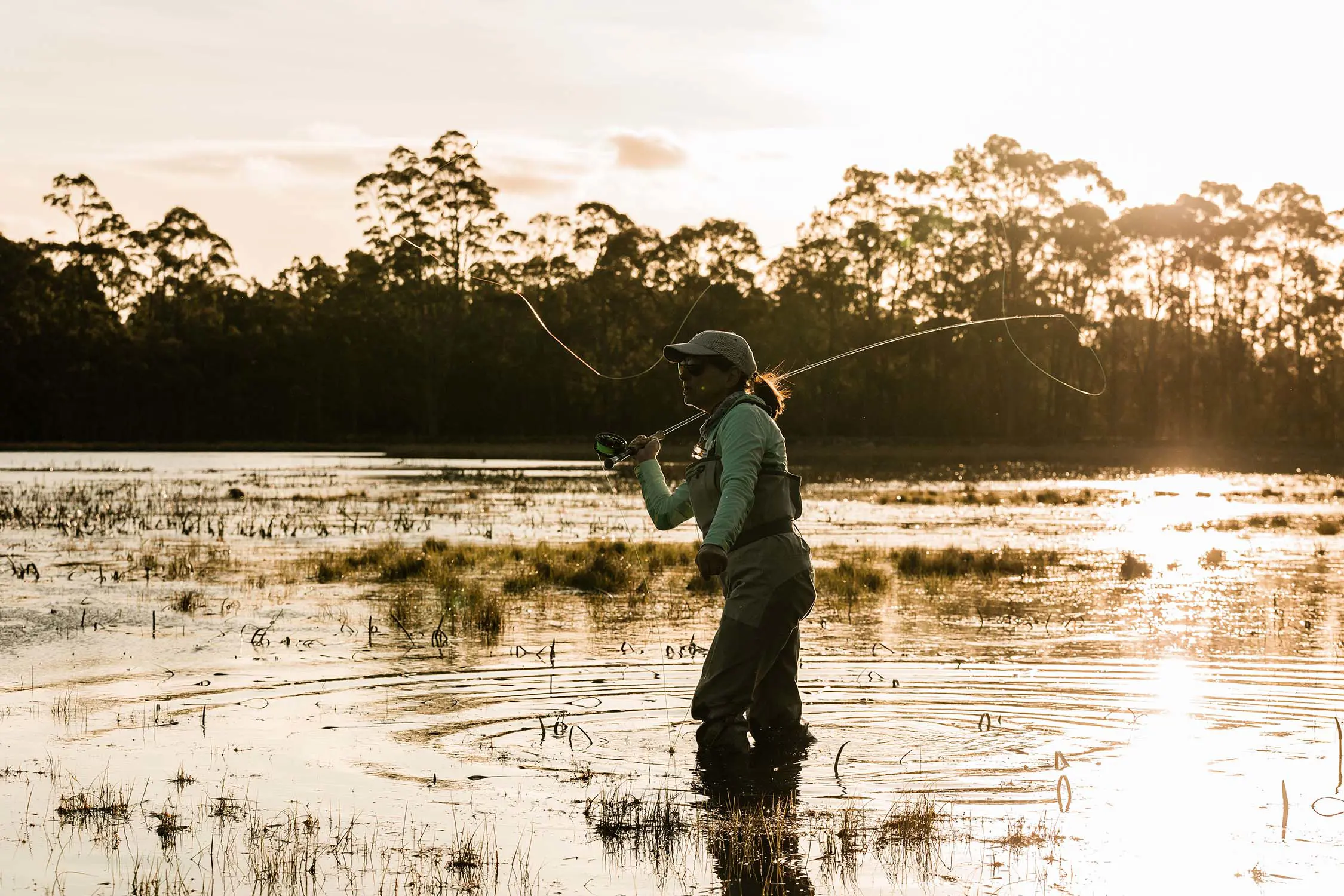 A woman in waders stands in the knee-deep water of a lake, a fishing line swinging from the rod over her shoulder.