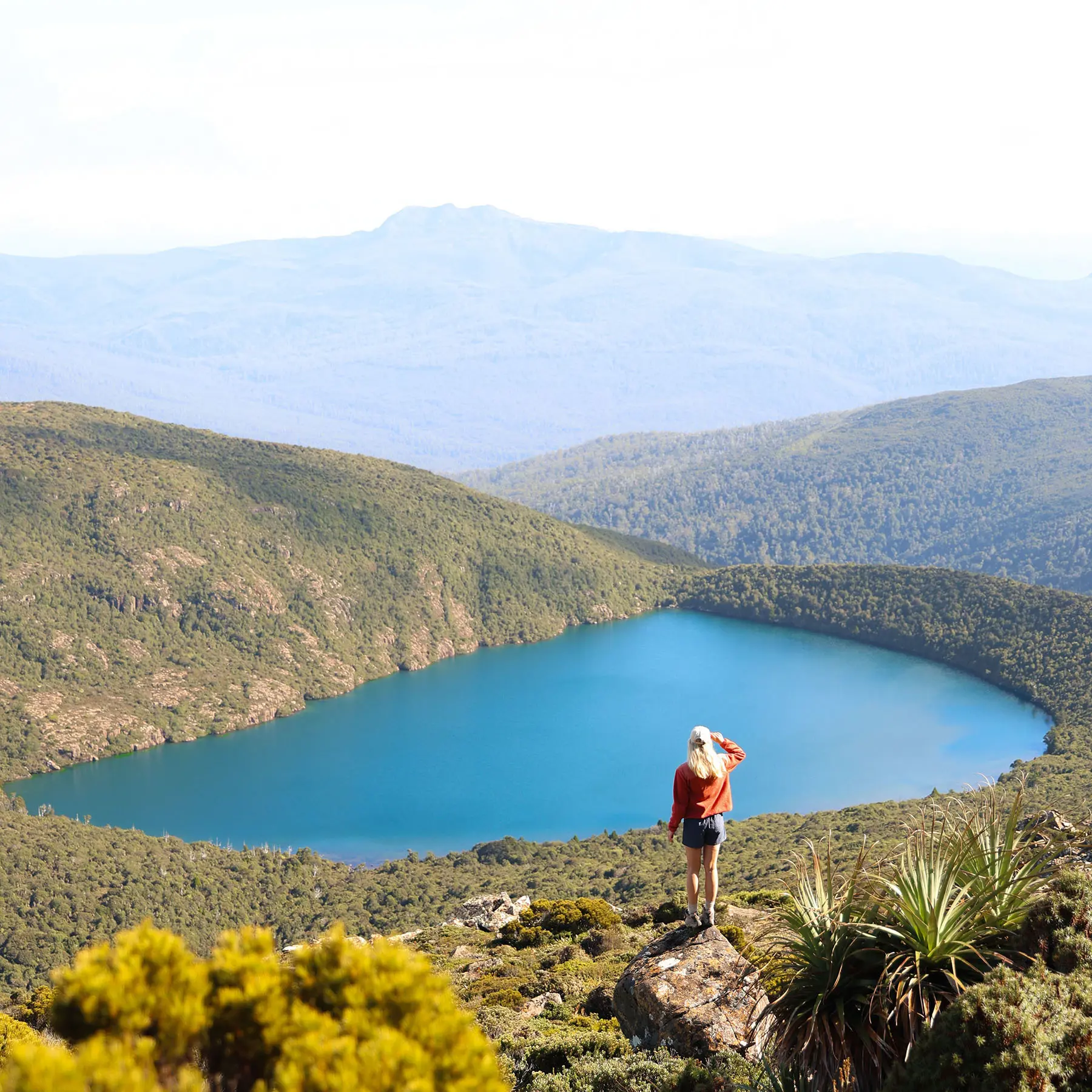 A hiker stands on a rock looking out over a beautiful still blue alpine lake, nestled in between rising mountains covered with dense green bush.