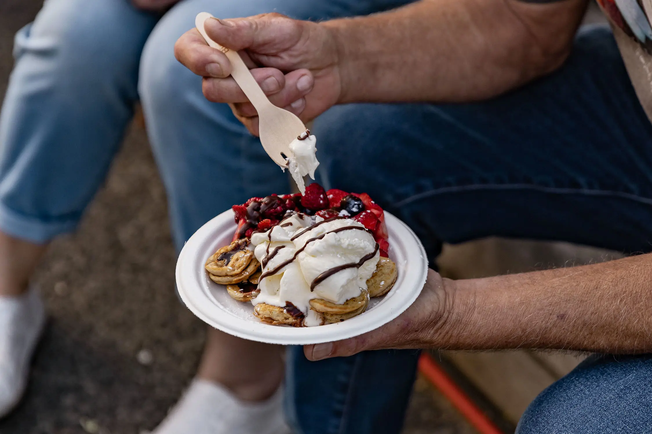 A person holds a paper plate at a food festival, filled with mini pancakes, fresh berries and icecream.
