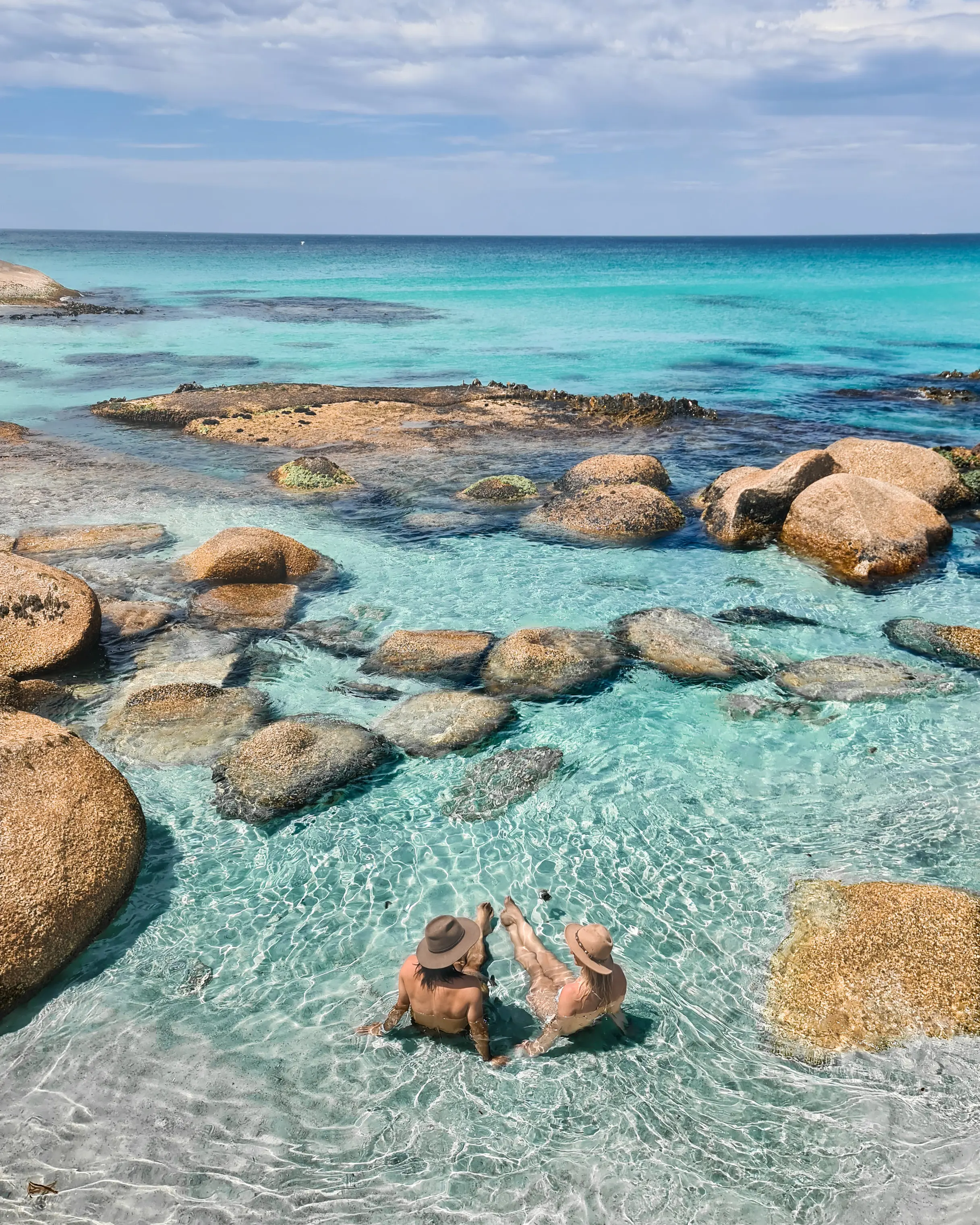 Two people relax in the crystal clear waters of a rocky beach inlet, looking out to the horizon.
