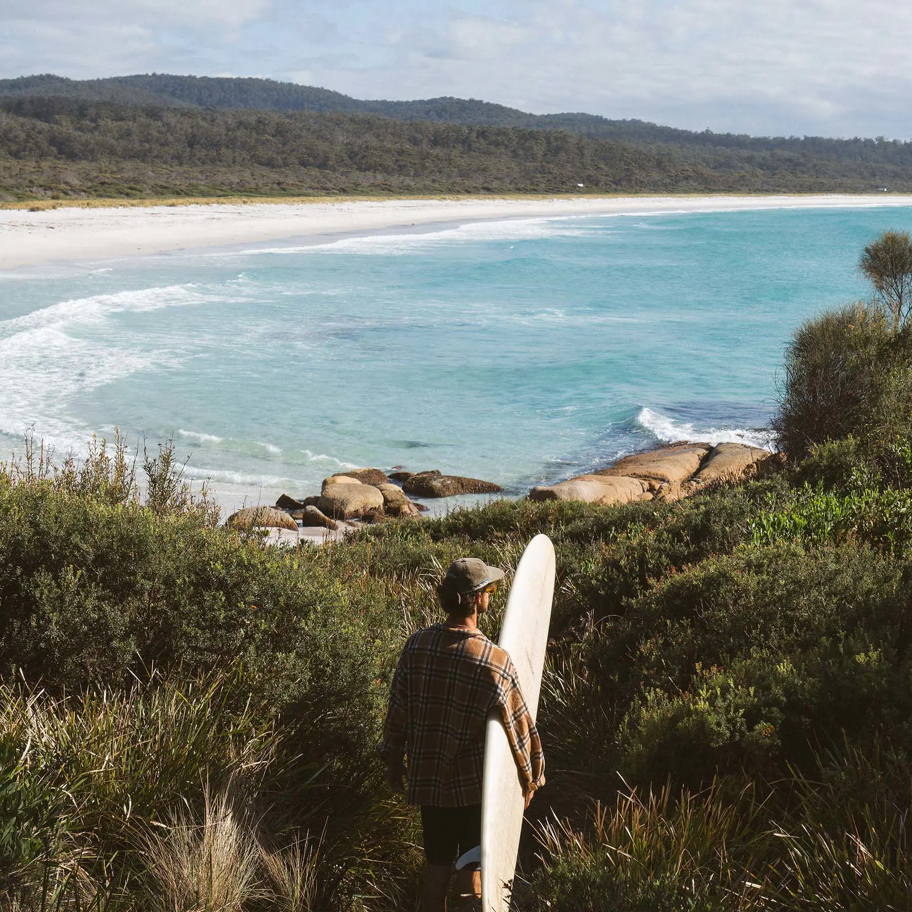 A person carrying a surfboard walks down a pathway towards a clear blue beach with waves rolling gently in.