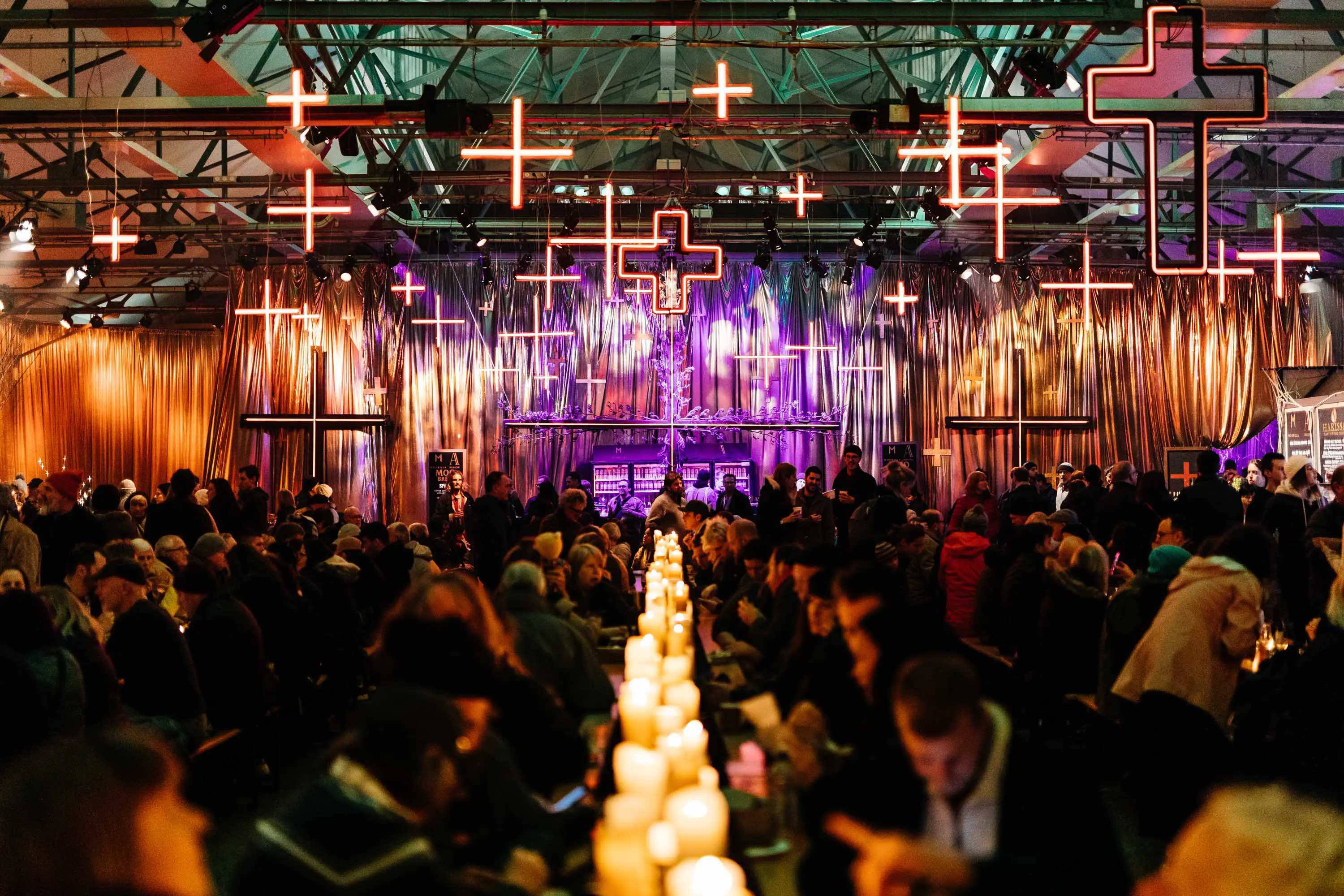 Hundreds of people sit at long benches, eating and drinking under decorations hanging form the eaves of a large high-ceiling wharf shed.