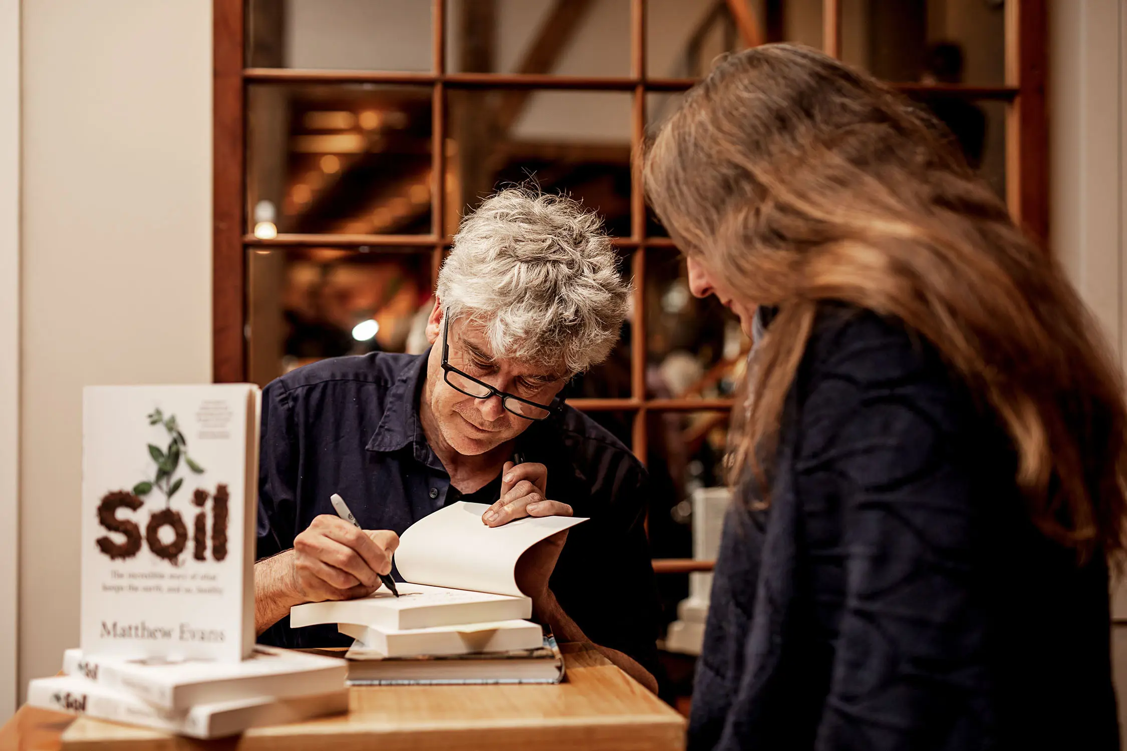 A man sits at a table leaning over a stack of books, signing the inside of the top book for a woman who is standing watching. 