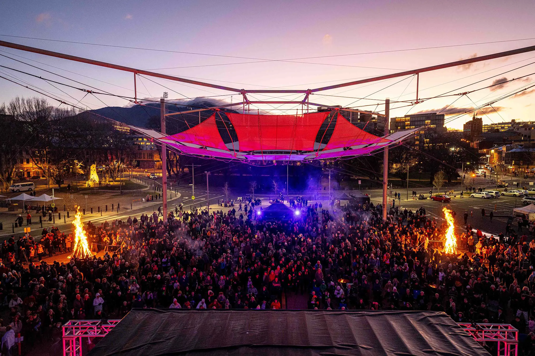 A large outdoor crowd gathered at dusk for a festival under a glowing canopy structure. Two bonfires at each end radiate warmth, and the twilight sky enhances the festive atmosphere.