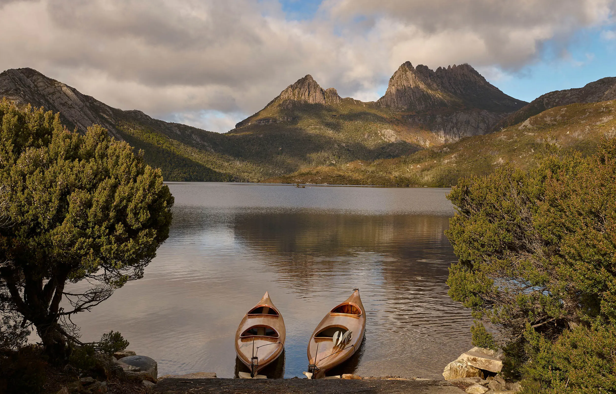 Two wooden kayaks rest on the shore of a pristine lake, with dramatic mountain peaks reflected in the calm water and surrounding bush.