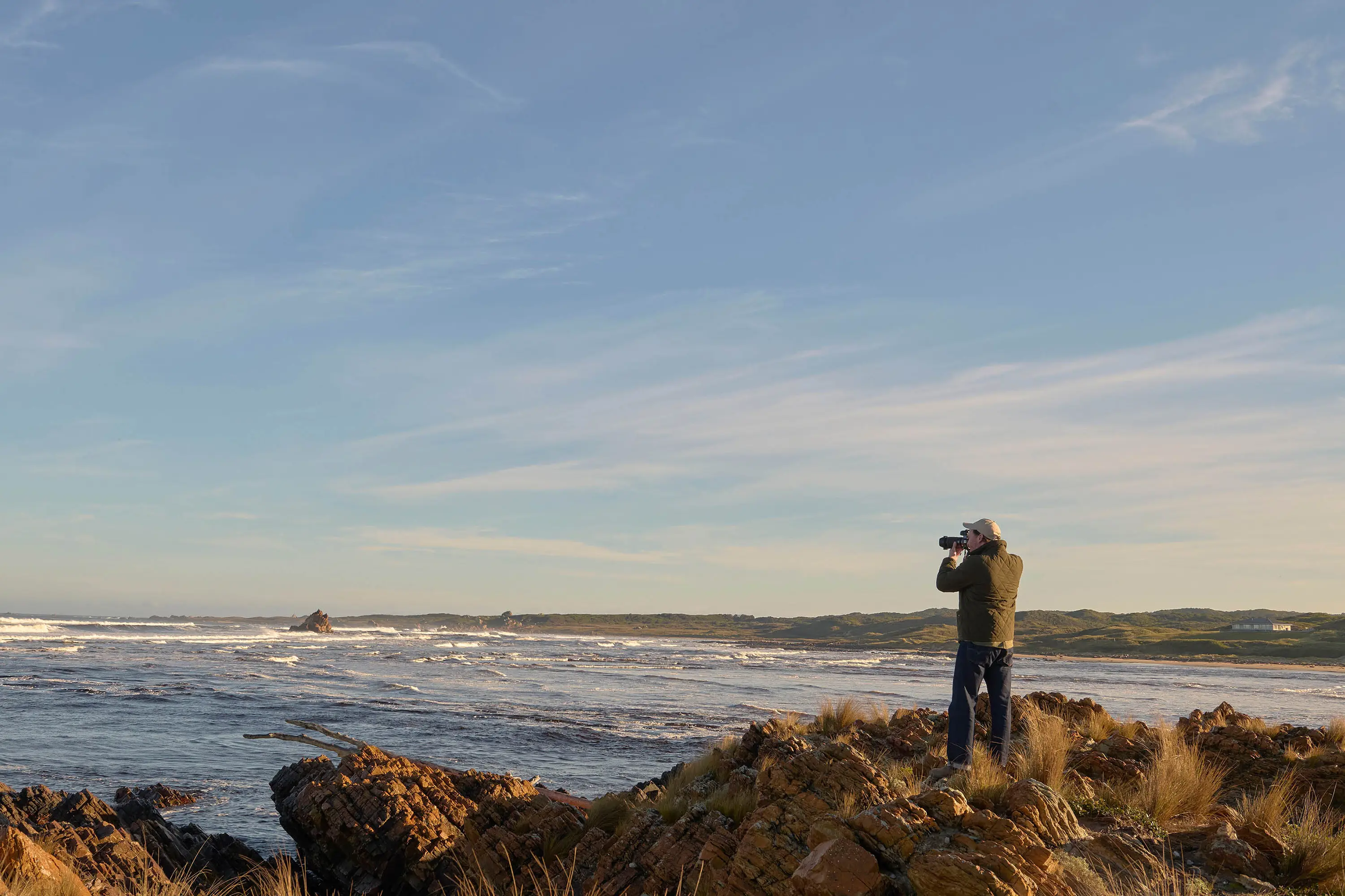 A man stands on the grassy rocks at the edge of the ocean, facing out to the horizon with a large camera raised to his face.