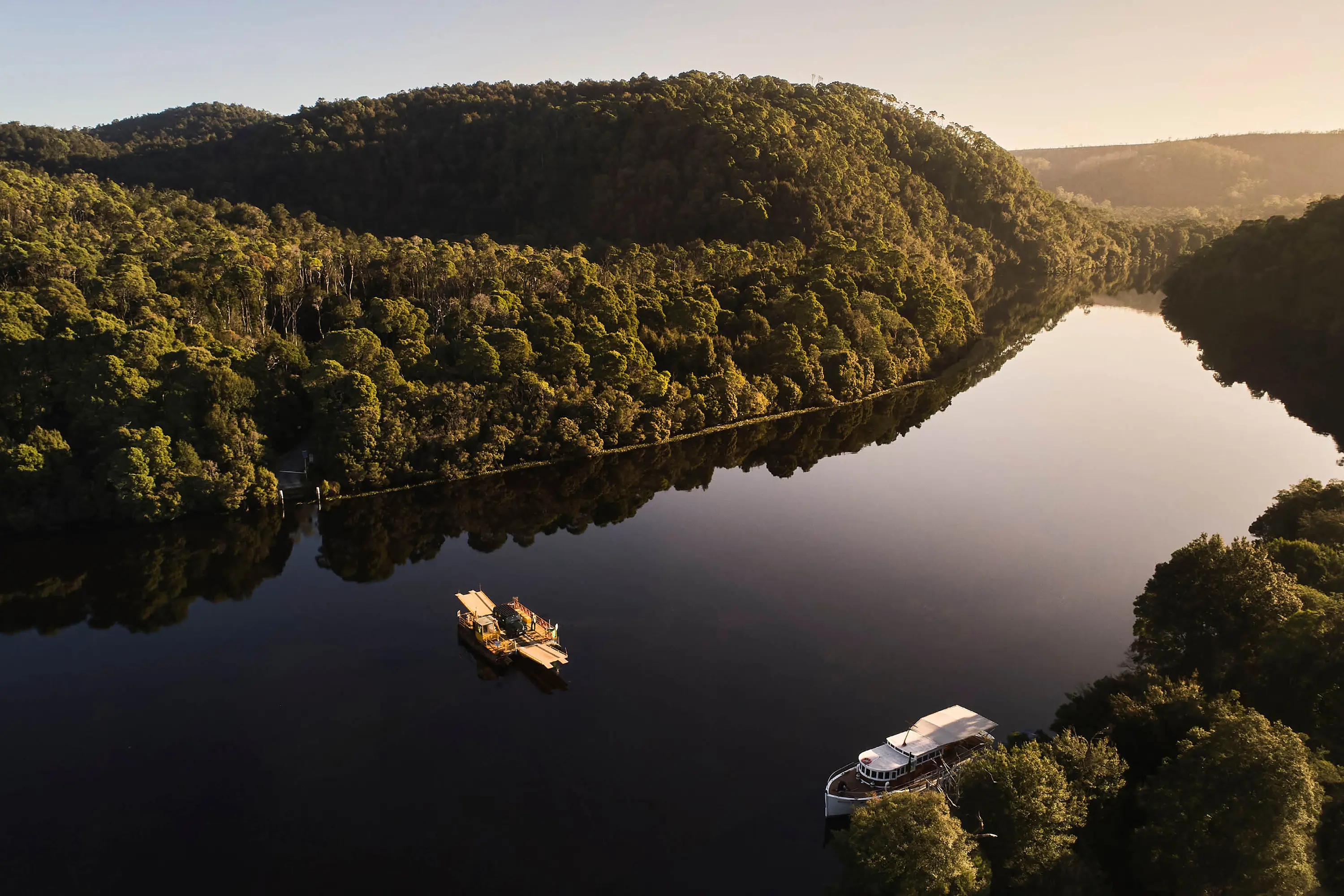 An aerial view of a still-as-glass river, banked on either side by deep densely green alpine rainforest. A small barge and a sleek boat are the only signs of human existence.