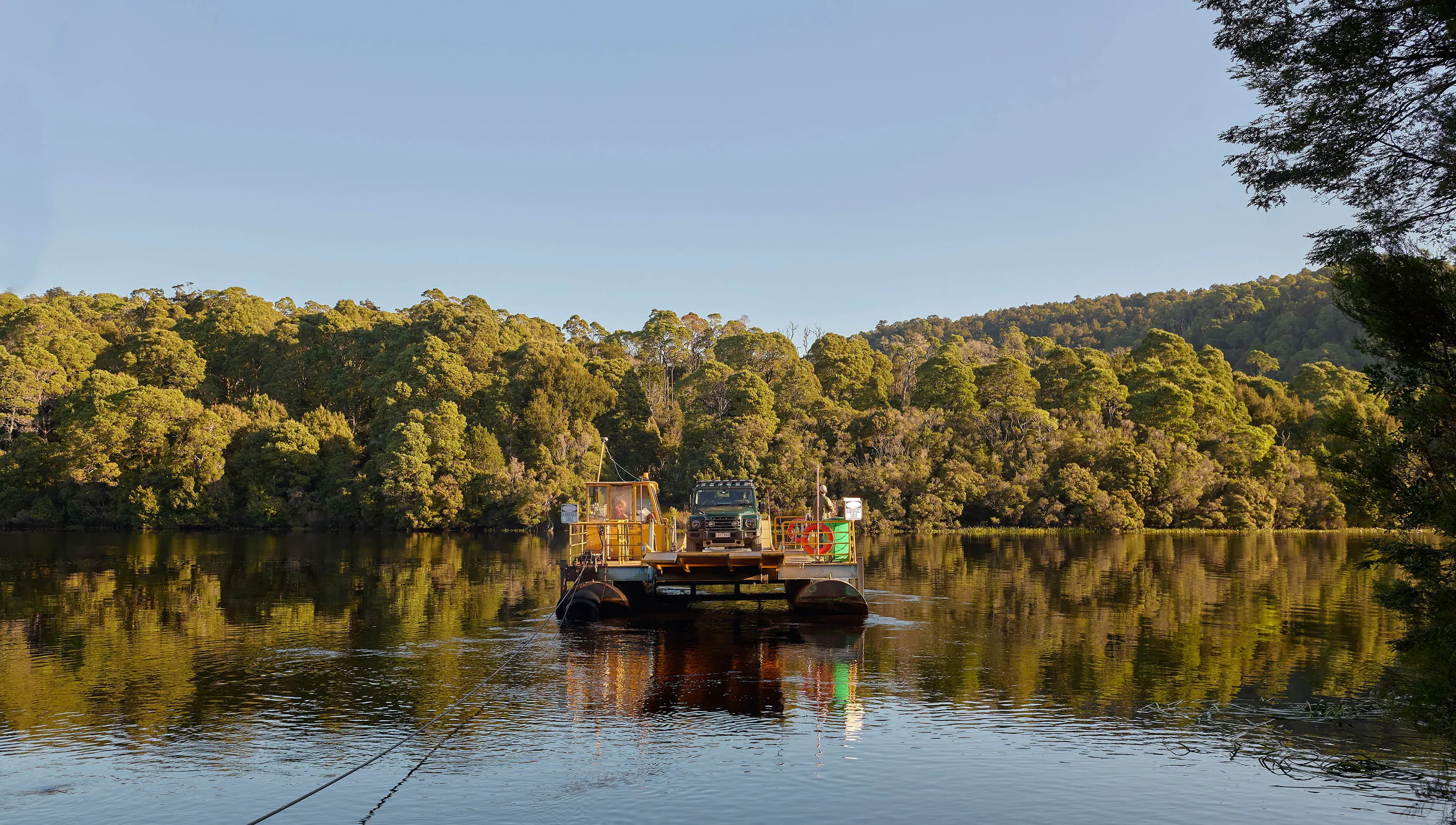 A small barge, loaded with a single car, floats on a river surrounded by dense native forest, which is reflected in the calm water.
