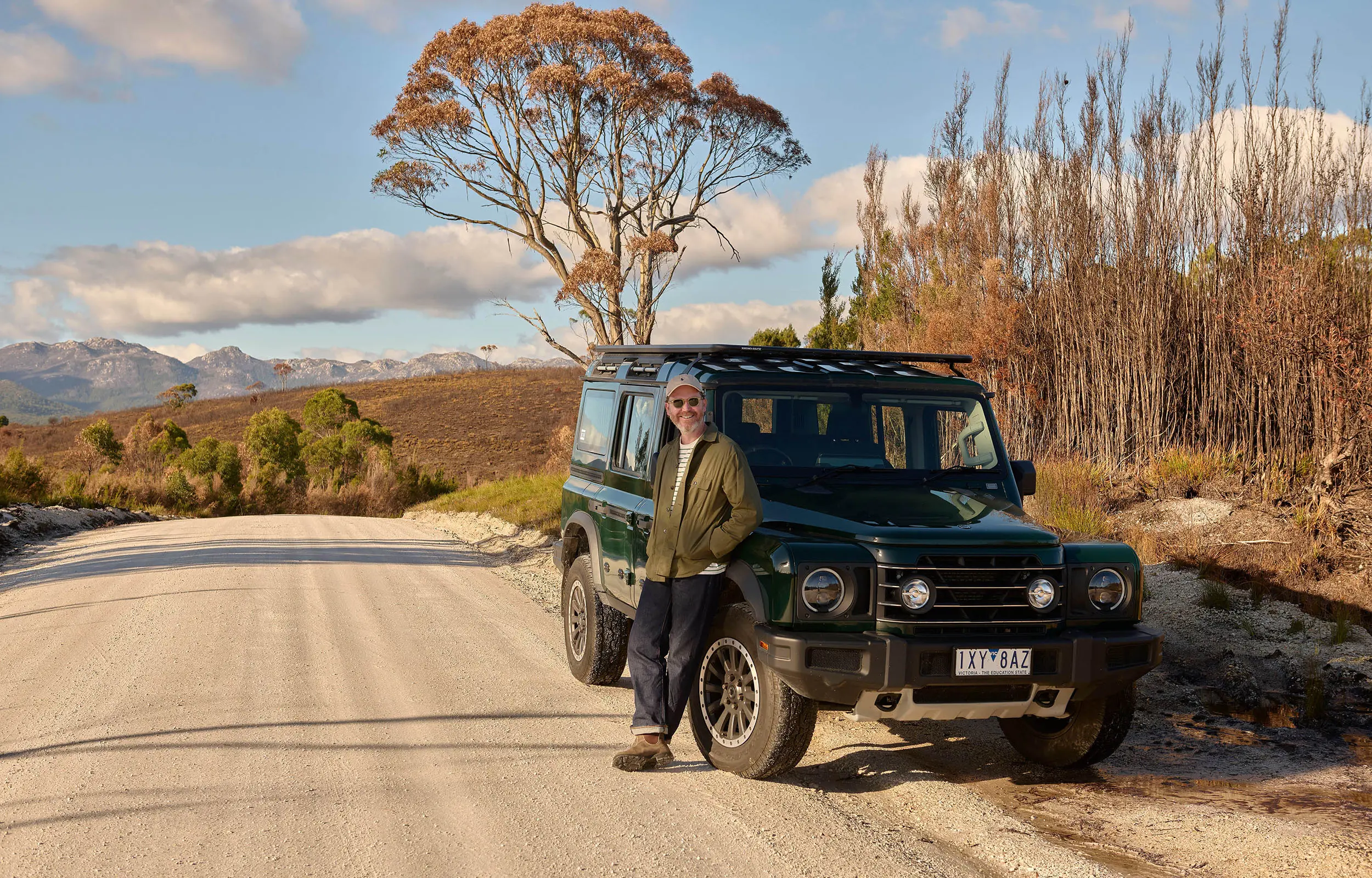 A person in a green jacket stands beside a black four-wheel-drive vehicle, parked on the shoulder of a gravel road. Mountains and alpine, scrubby vegetation are visible in the background.