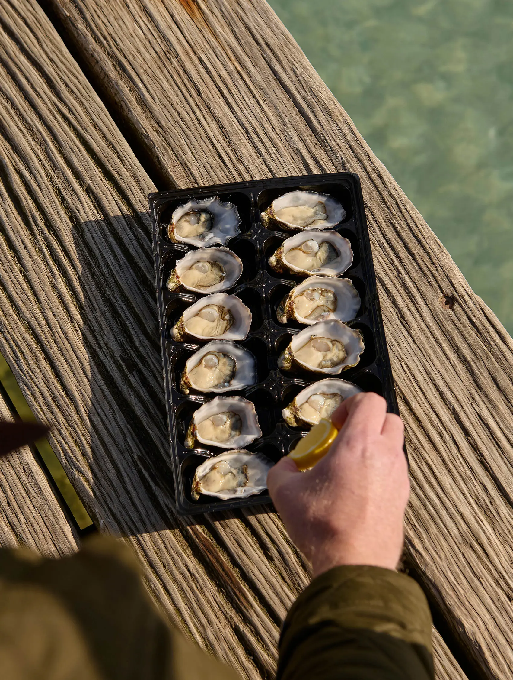 A person's hand squeezes fresh lemon over a black tray of a dozen oysters, sitting on a weathered wooden surface beside the water.