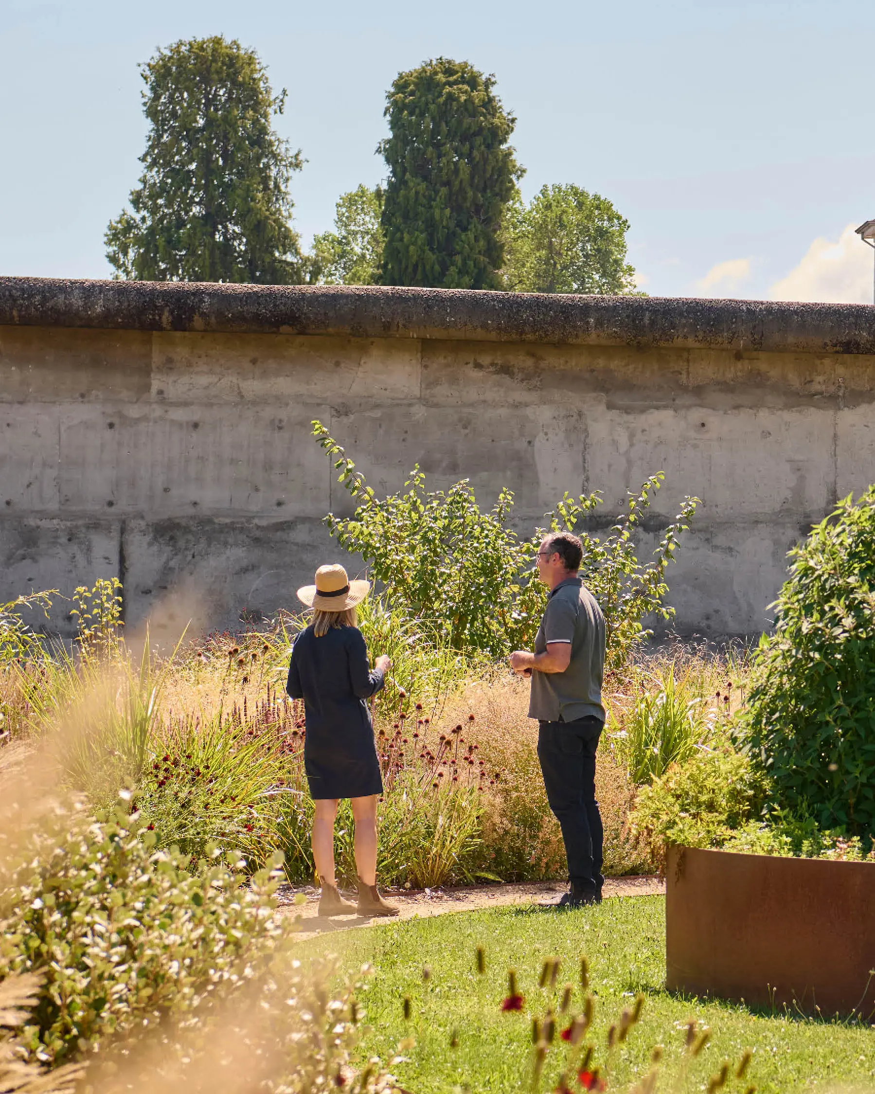 Two people stand in a walled garden, with the thick grasses and bushes contrasting with manicured lawn.