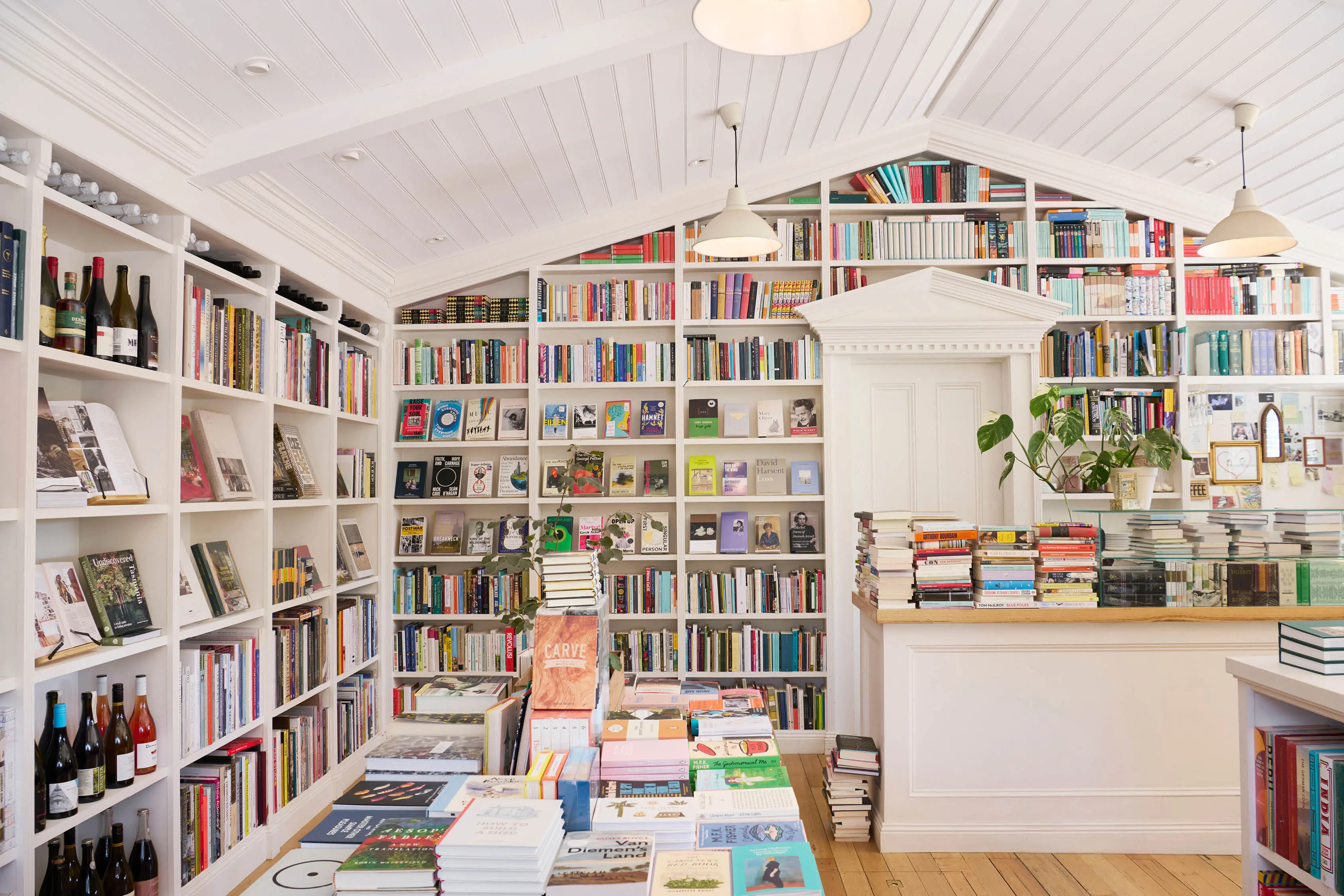 A bookshop with floor-to-ceiling white shelves stacked full of colourful books. Central tables and counters are piled high, and the space is light-filled and airy.