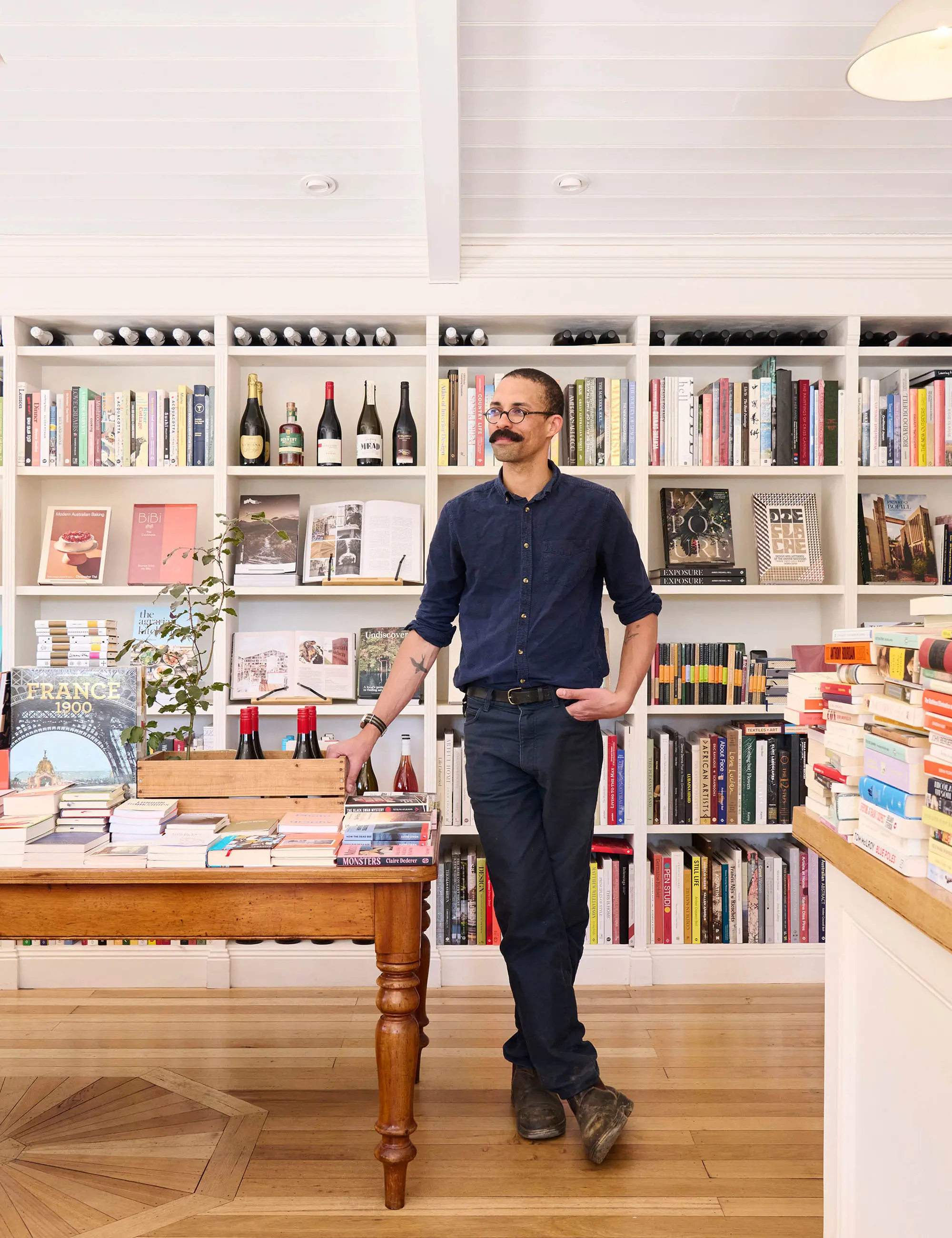 A man stands inside a bookshop, one hand in his jeans pocket, leaning against a vintage wooden table filled with an array of colourful books.