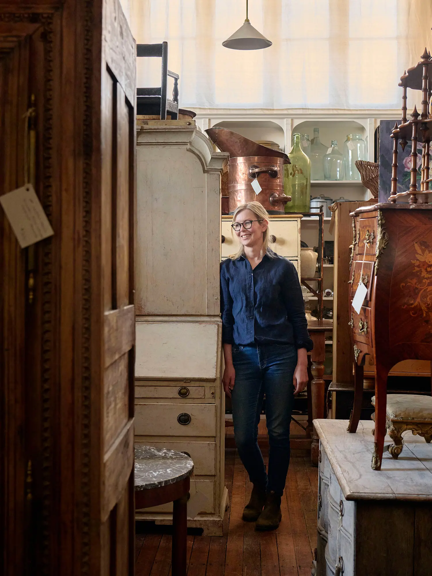 A woman in a blue shirt and jeans stands amongst an eclectic collection of antique and vintage furniture, stacked high inside a shop.
