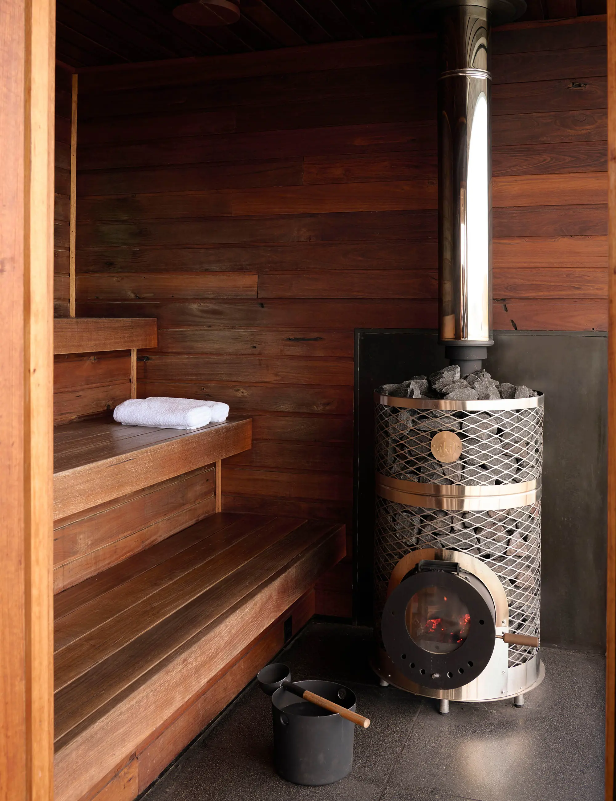 A small, wood-clad sauna room, with stepped seating up one side, and a sleek sauna stove filled with rocks.