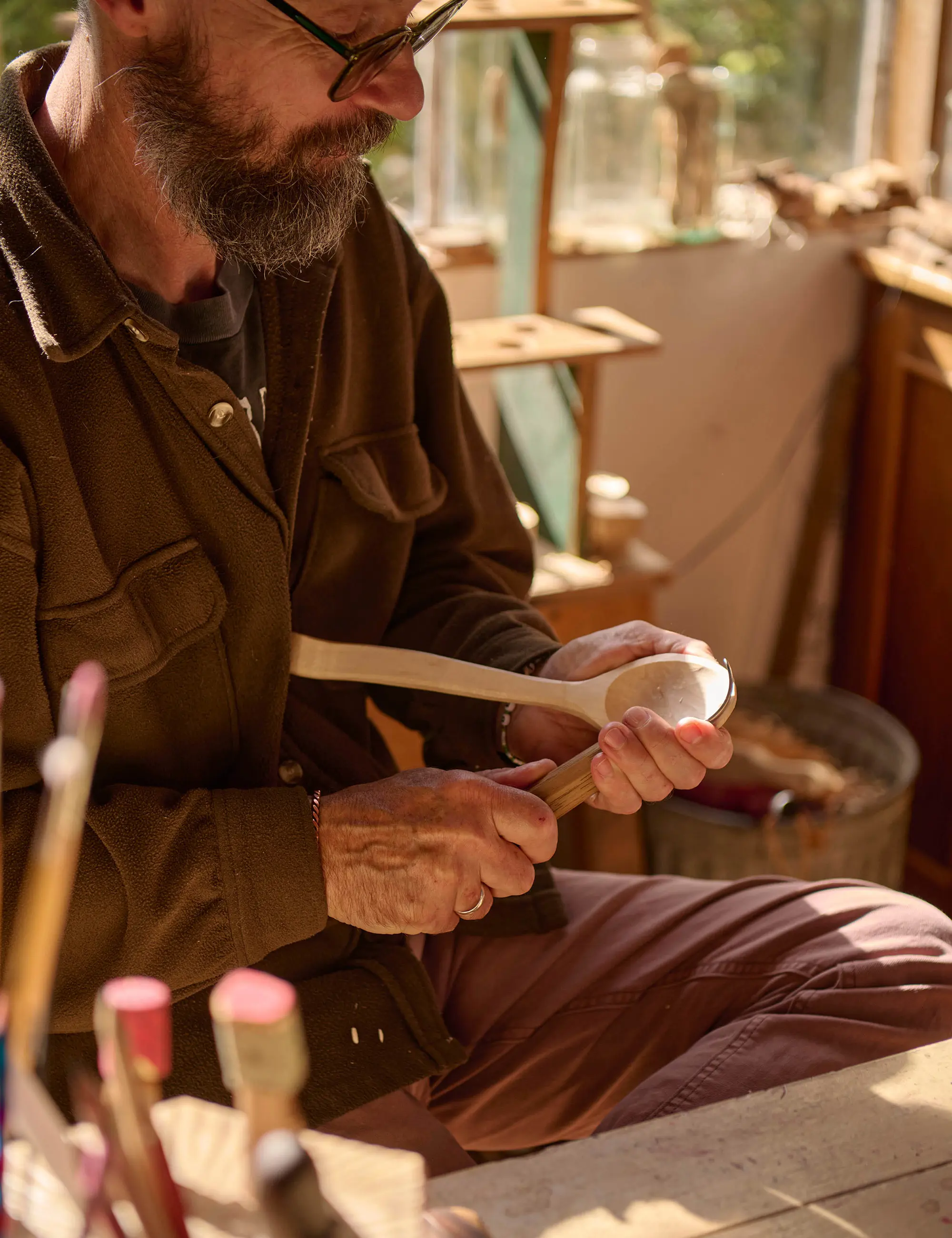 A bearded man sits in a light-filled workshop, carving the bowl of a wooden spoon using a hook-shaped tool.