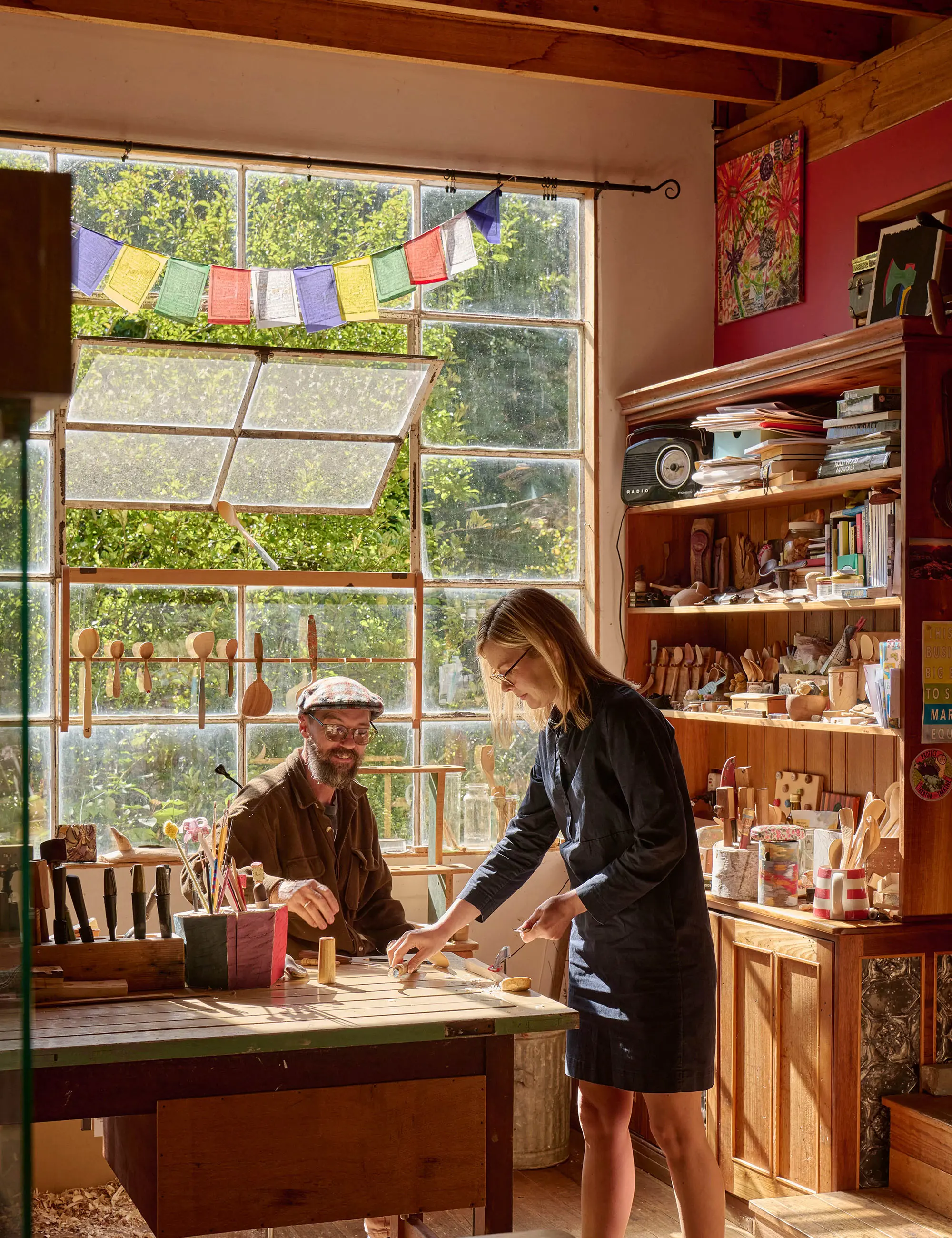 A man and woman in a light-filled woodworking studio, surrounded by carved spoons and tools. The light streams in the wide antique window behind them.