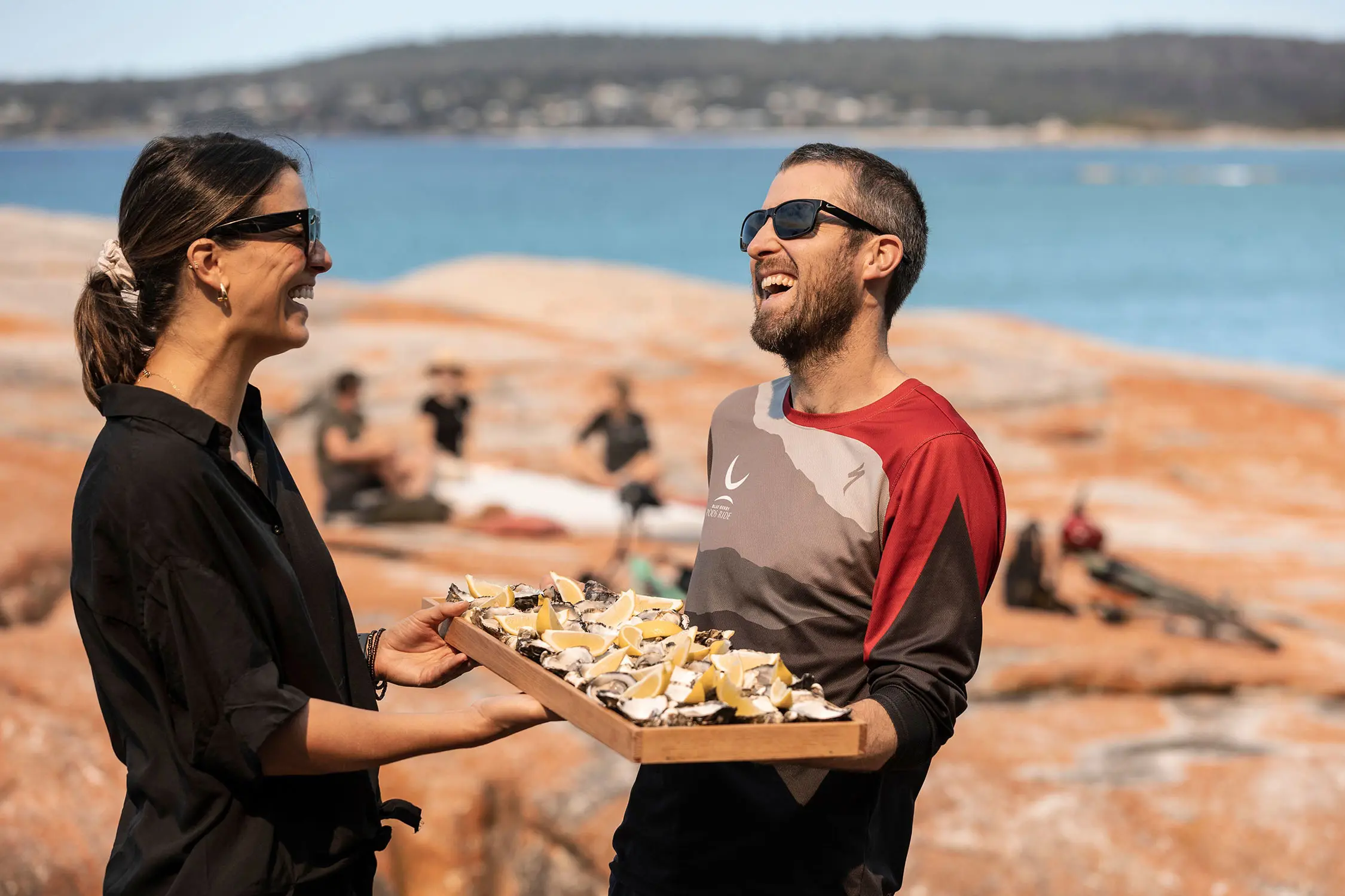 Two people stand on the orange lichen-covered rocks at a beach, holding a tray of oysters topped with lemon wedges.