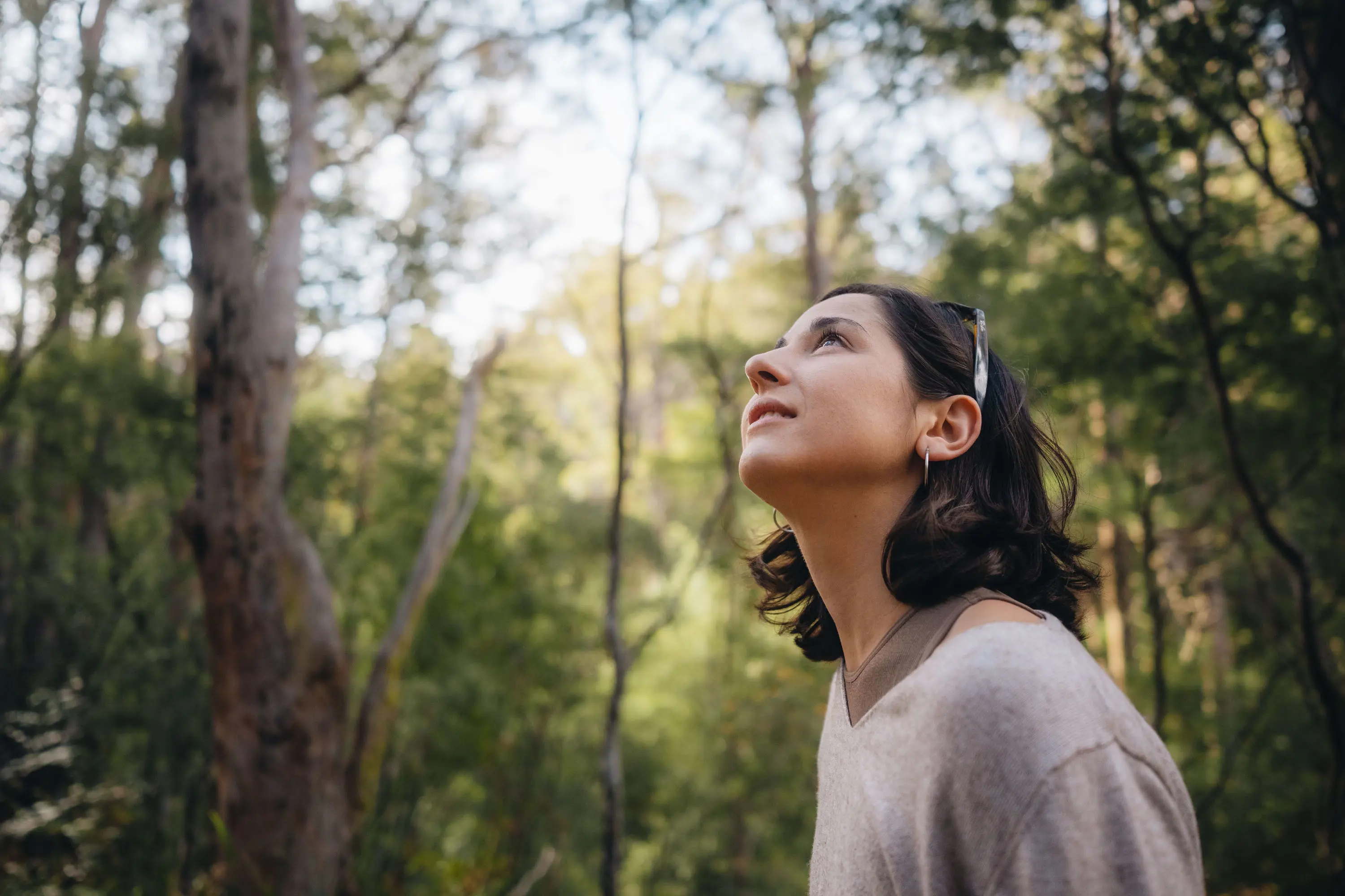 A woman stands in a forest setting, wearing a cream jumper. She is gazing up into the canopy above her, a peaceful awed expression on her face.