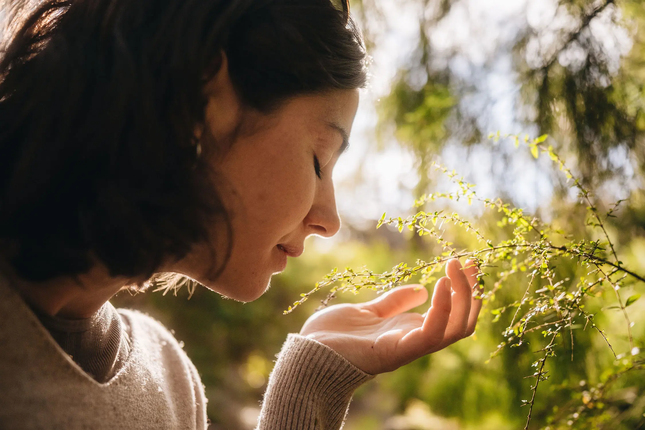 A close up of a woman as she leans forward and lifts a delicate branch of foliage up to her face.