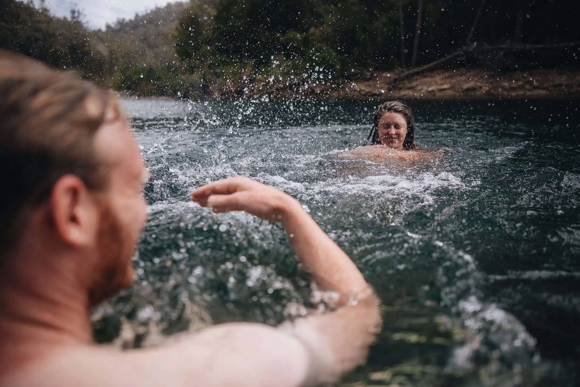 Two people swim in the dark waters of a lake, one splashing water onto the other as they laugh.