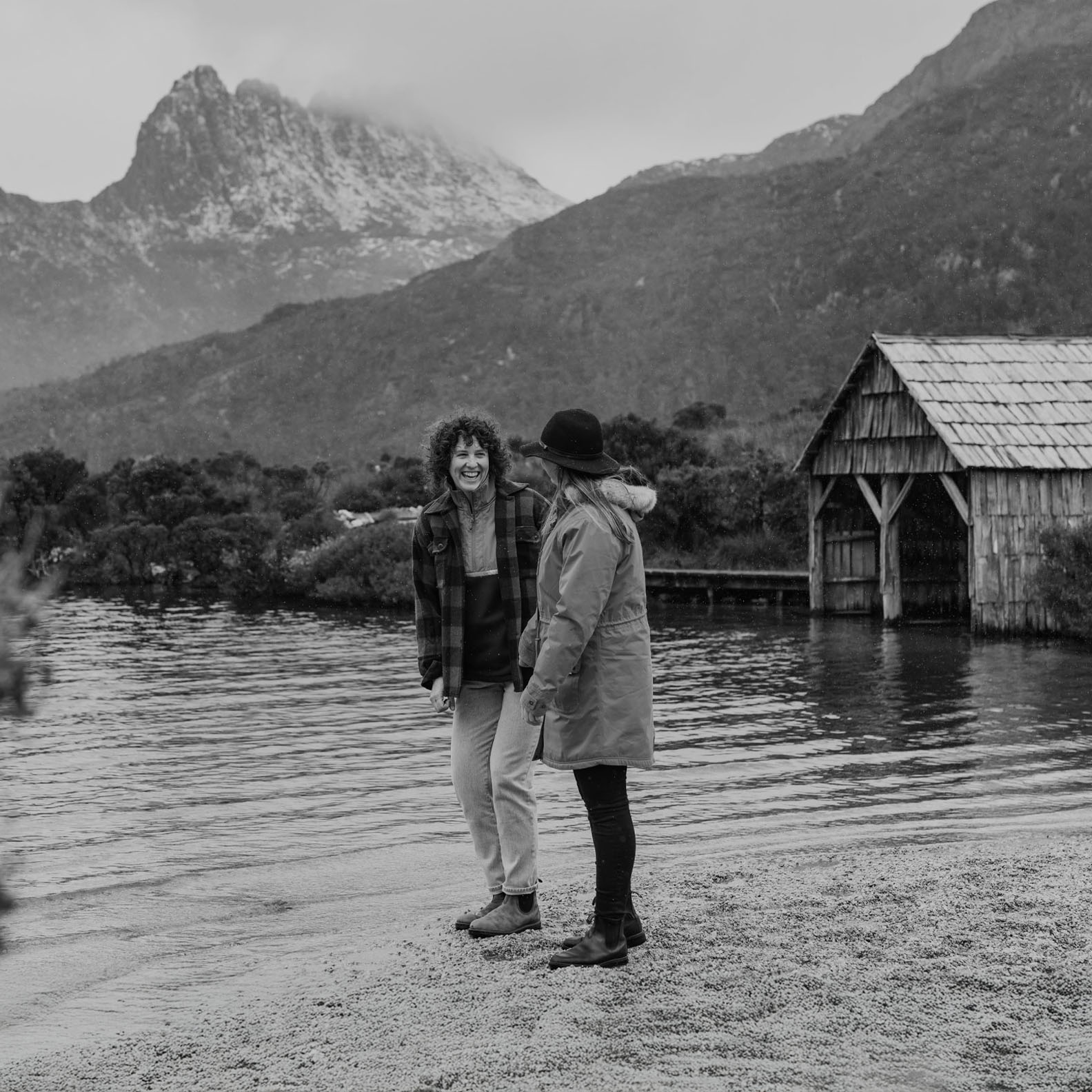 Two women are standing near the edge of a calm lake, laughing together. One is wearing a plaid jacket, and the other is dressed in a warm coat and hat. Behind them, mist-covered mountains rise dramatically into the sky, and a rustic wooden boat shed sits at the water's edge. The scene exudes a sense of joy and connection in a beautiful natural environment